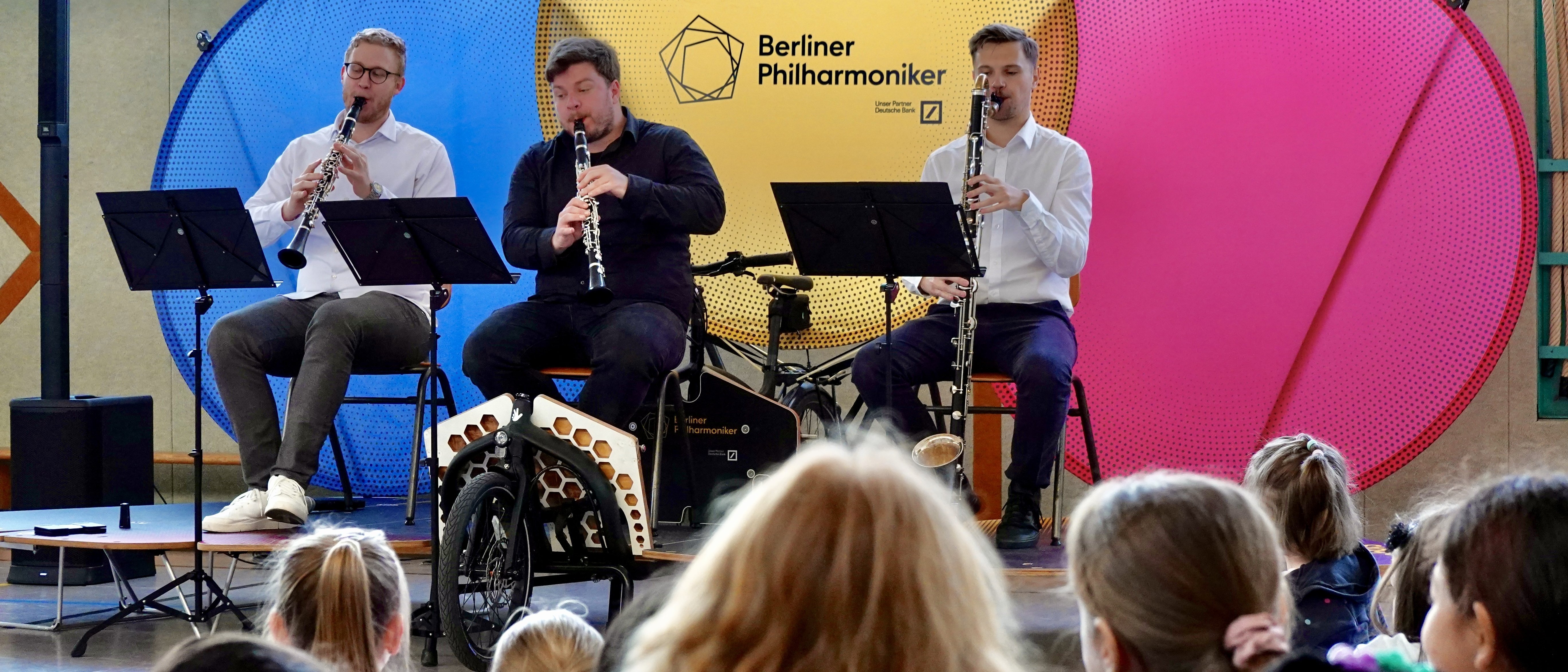 Three musicians play clarinets on a small stage in front of an audience of children, with colorful circular backdrops and a “Berliner Philharmoniker” sign behind them.