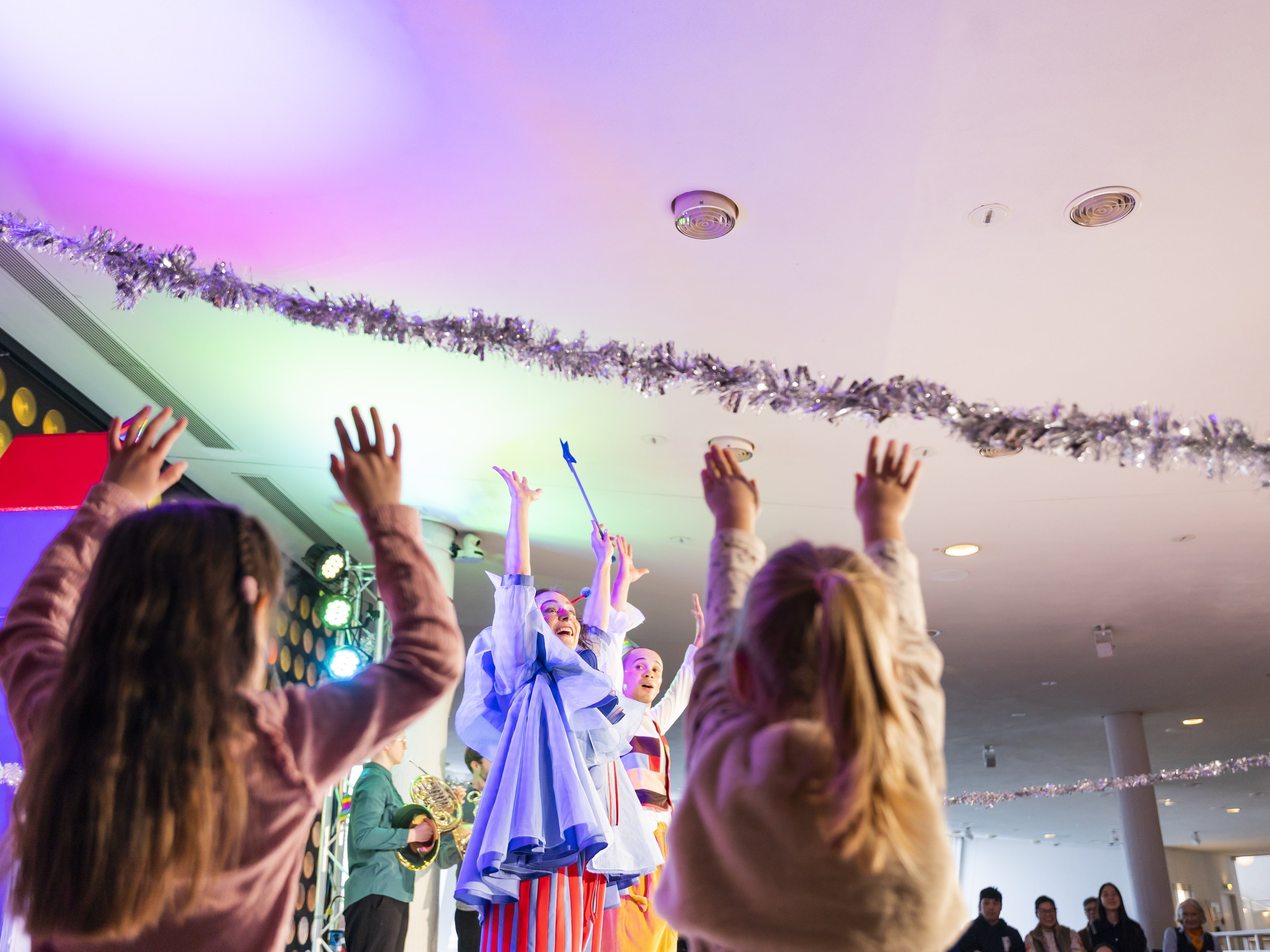 Kinder mit erhobenen Armen beobachten Darsteller in bunten Kostümen auf einer hell erleuchteten Bühne, von deren Decke festliche Dekorationen hängen. Die Atmosphäre wirkt fröhlich und lebendig.