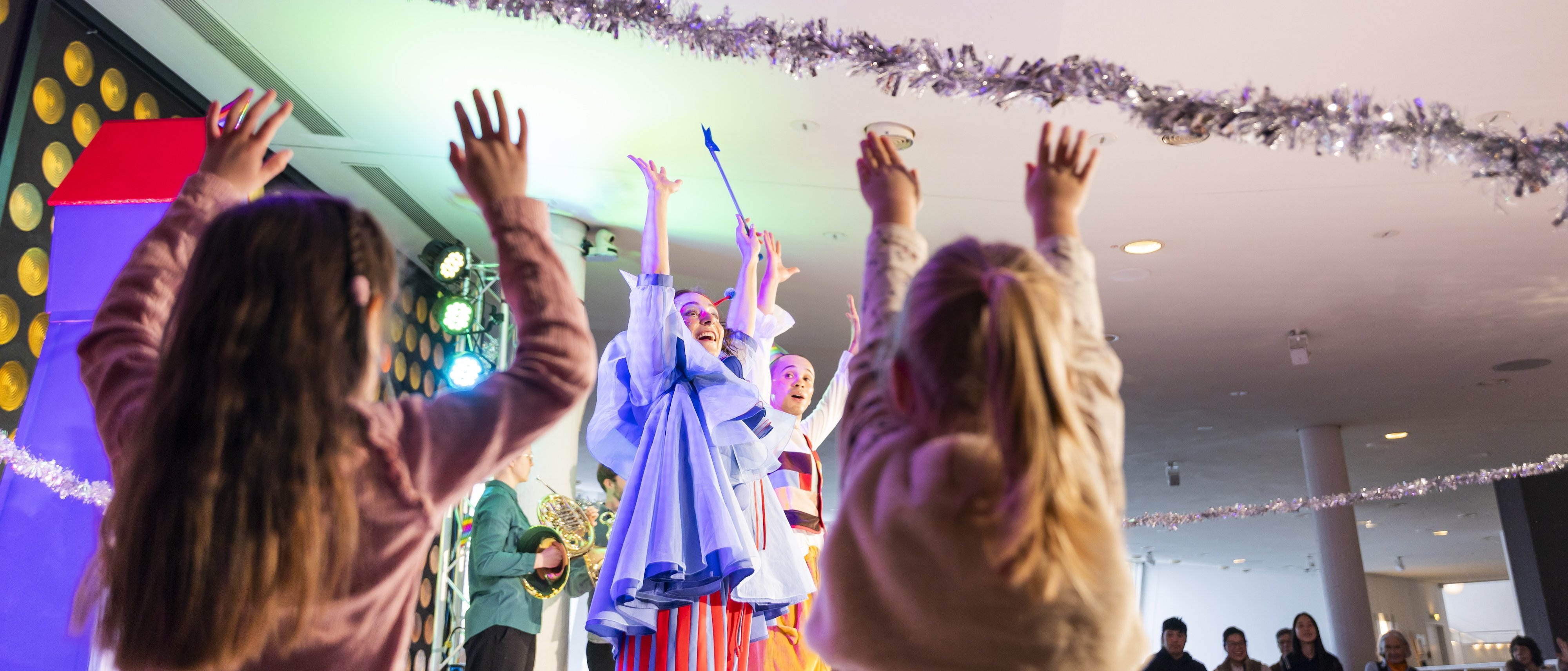 Kinder mit erhobenen Armen beobachten Darsteller in bunten Kostümen auf einer hell erleuchteten Bühne, von deren Decke festliche Dekorationen hängen. Die Atmosphäre wirkt fröhlich und lebendig.