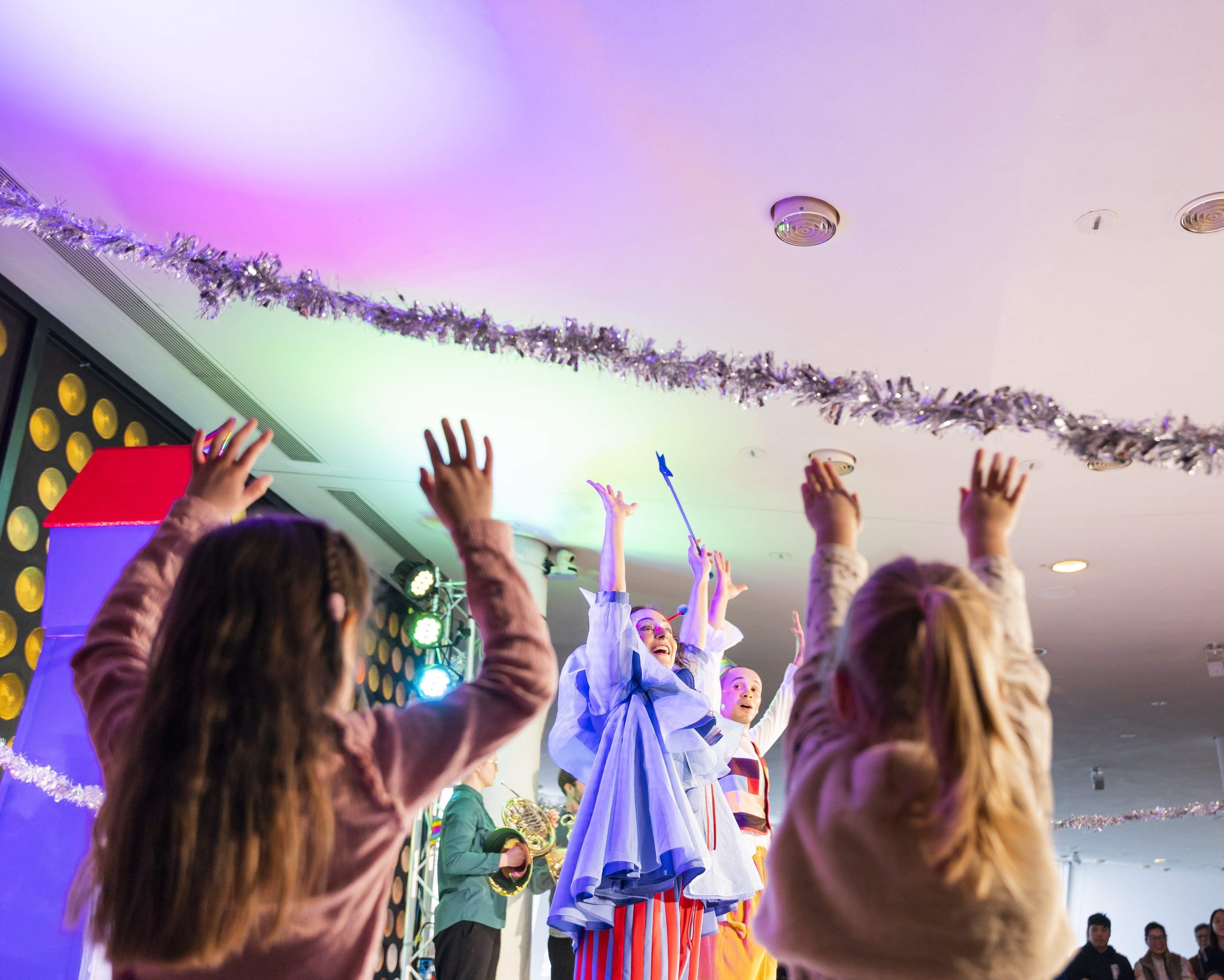 Kinder mit erhobenen Armen beobachten Darsteller in bunten Kostümen auf einer hell erleuchteten Bühne, von deren Decke festliche Dekorationen hängen. Die Atmosphäre wirkt fröhlich und lebendig.