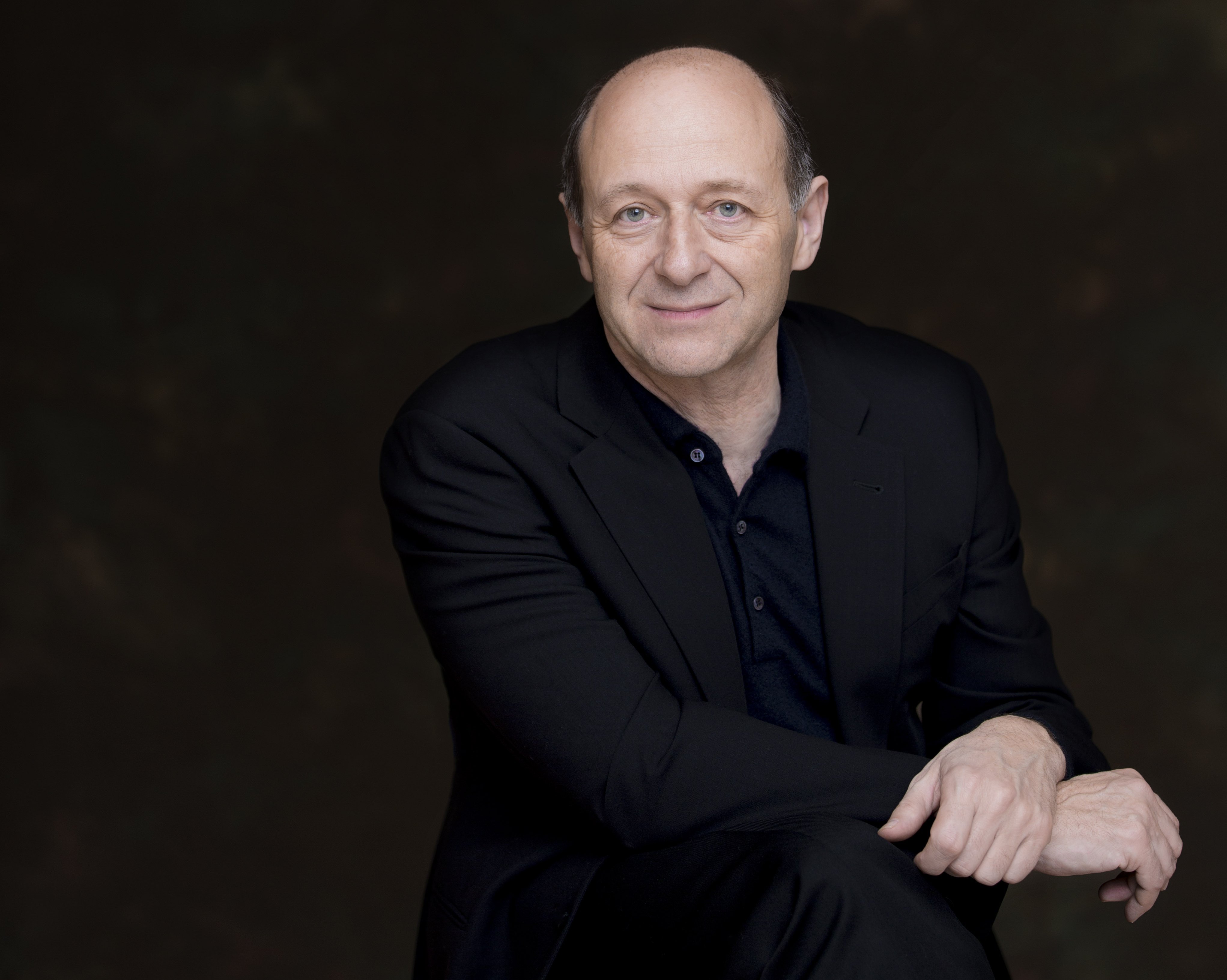 Iván Fischer mit zurückweichendem Haaransatz, der einen schwarzen Anzug trägt, sitzt mit einem Arm auf seinem Knie vor einem dunklen, leicht verschwommenen Hintergrund. Er blickt in die Kamera und lächelt leicht.