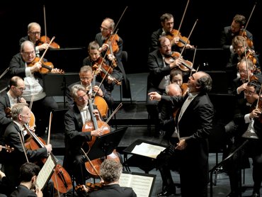 Ein Dirigent leitet ein Orchester von Musikern, die auf der Bühne Streichinstrumente spielen. Die Musiker, die förmlich in schwarze Anzüge gekleidet sind, konzentrieren sich im Bühnenlicht auf ihre Notenblätter und Instrumente.