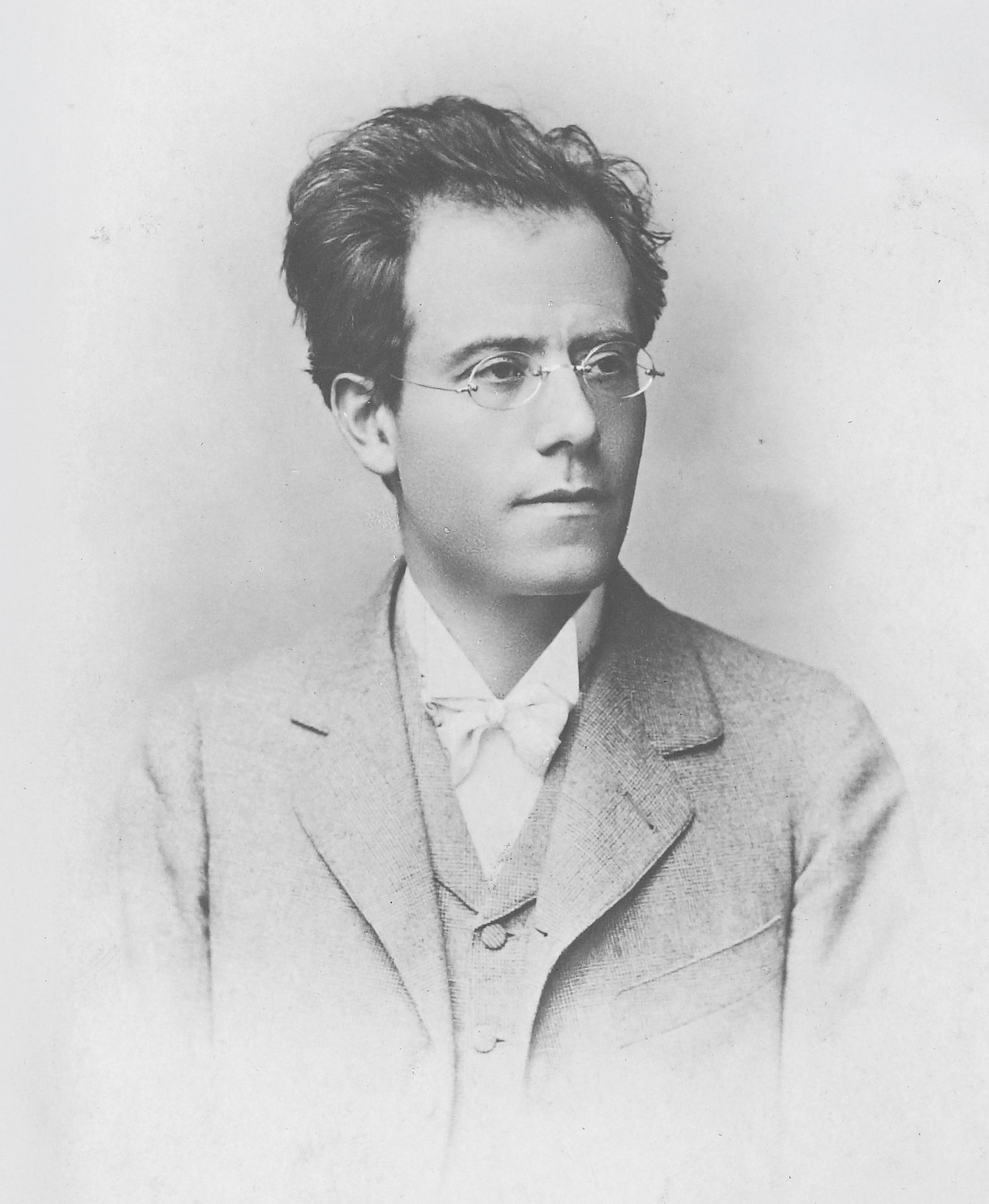 Black-and-white portrait of a young man with glasses, wearing a suit and bow tie, looking slightly to the side with a neutral expression. The background is plain and light-colored.