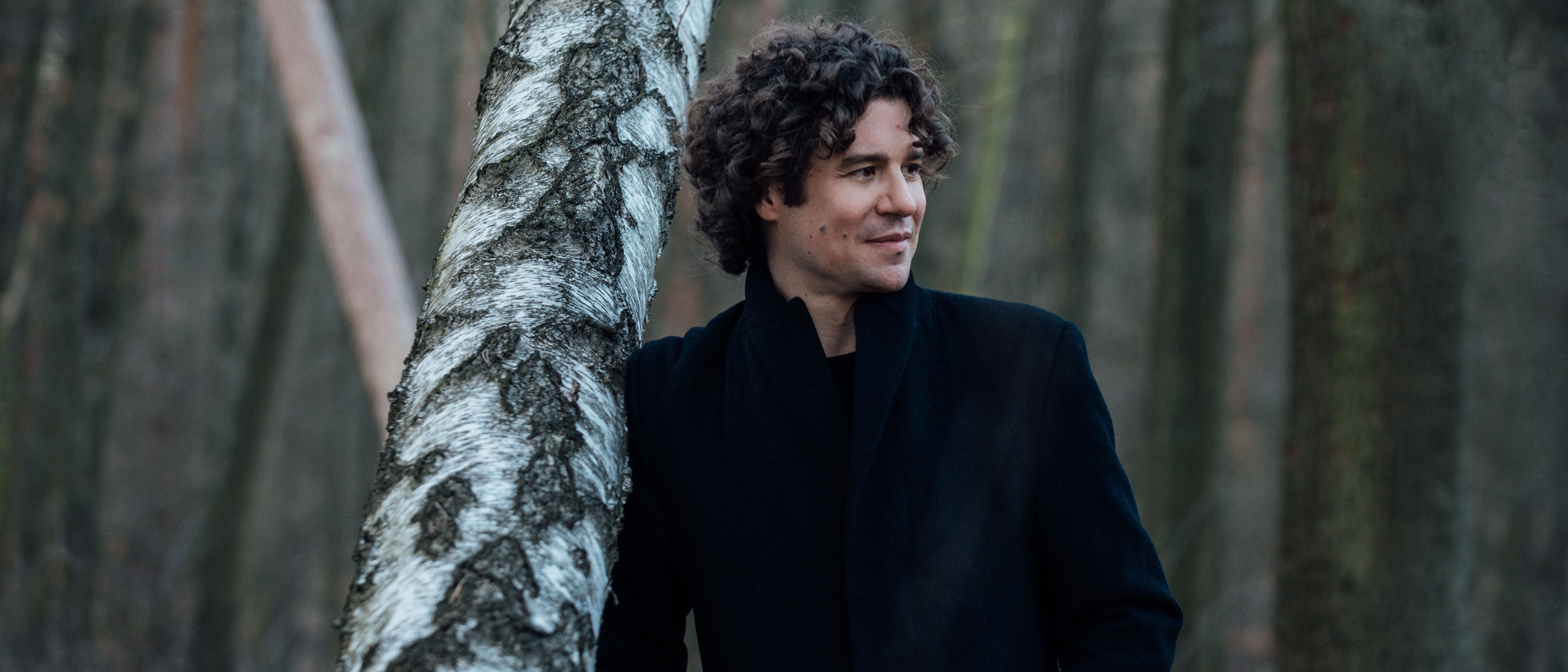 Robin Ticciati lehnt im Mantel an einem Baum im Wald