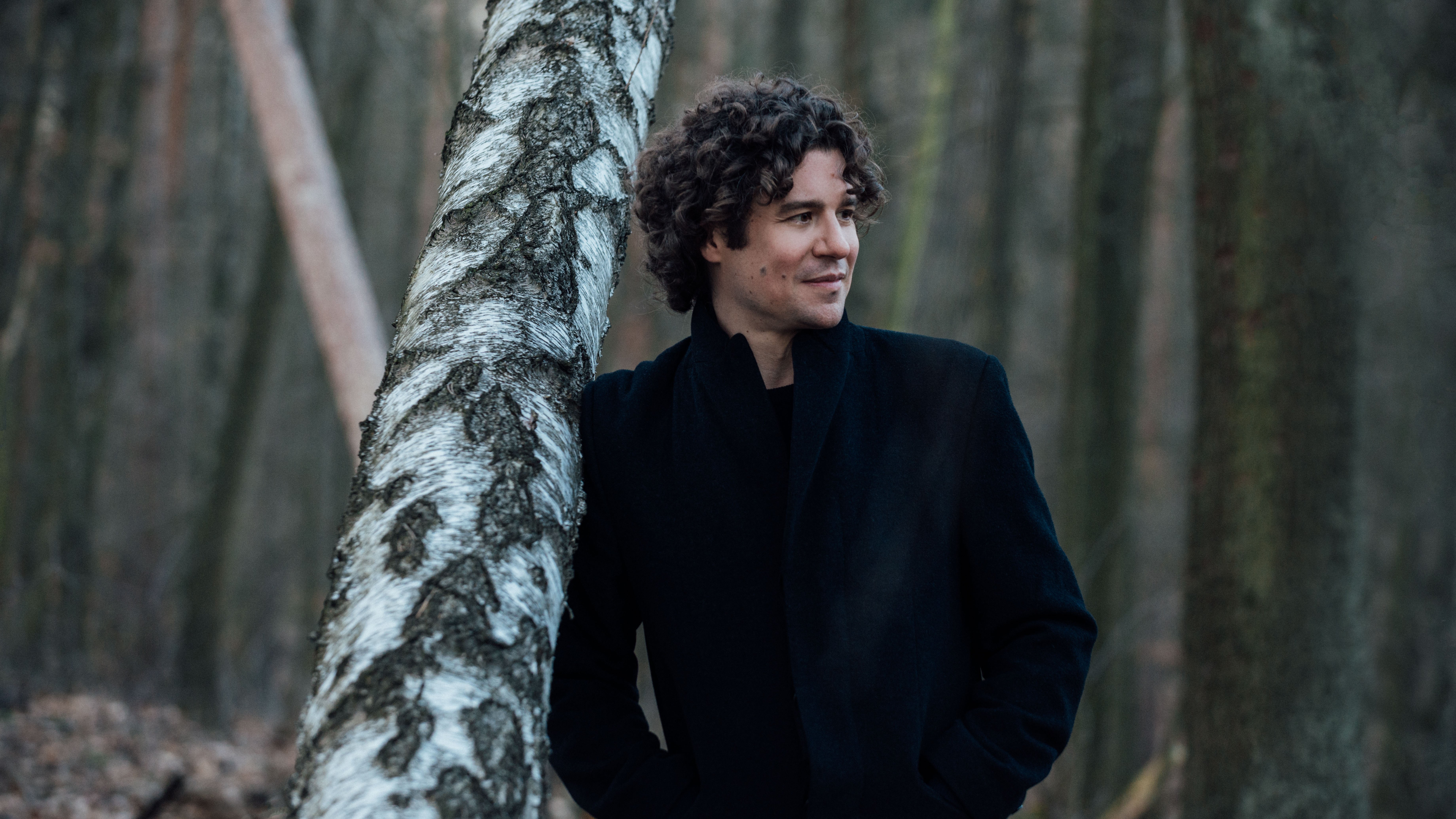 Robin Ticciati leans against a tree in the forest in his coat
