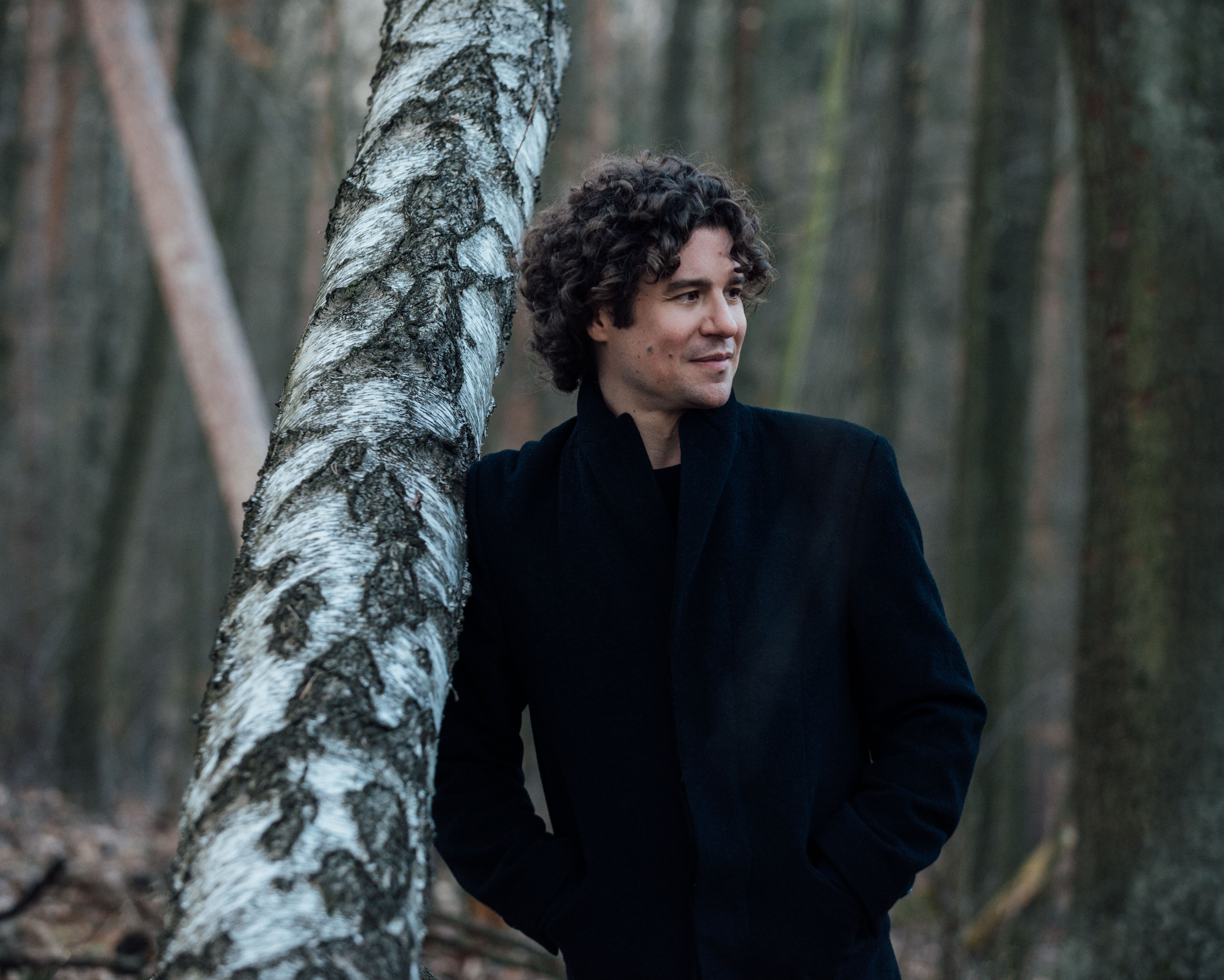 Robin Ticciati lehnt im Mantel an einem Baum im Wald