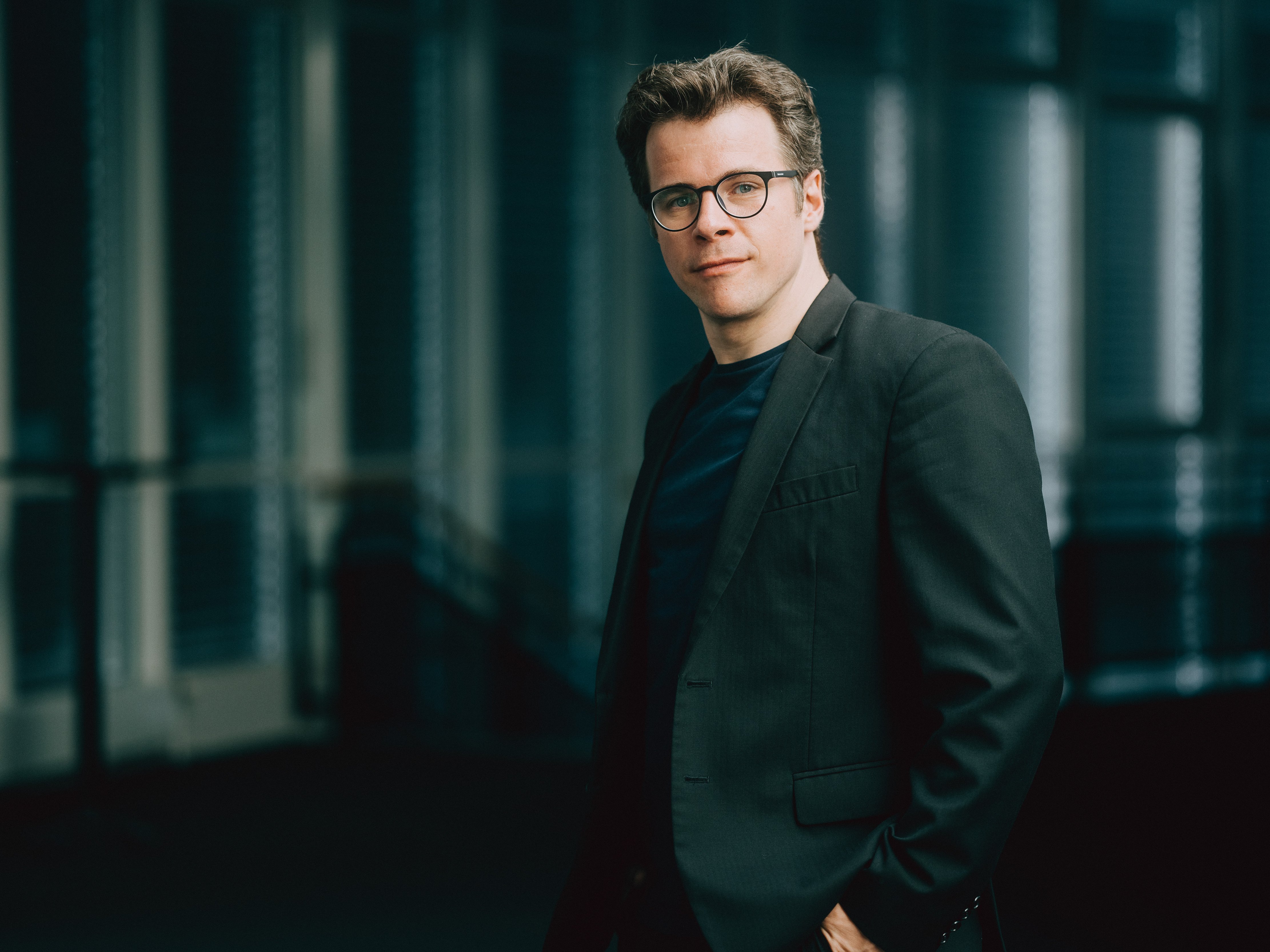 Jakub Hrůša mit Brille und dunklem Anzug blickt mit einem leichten Lächeln in die Kamera. Er steht in einer Innenumgebung mit sanftem Licht und unscharfem Hintergrund.