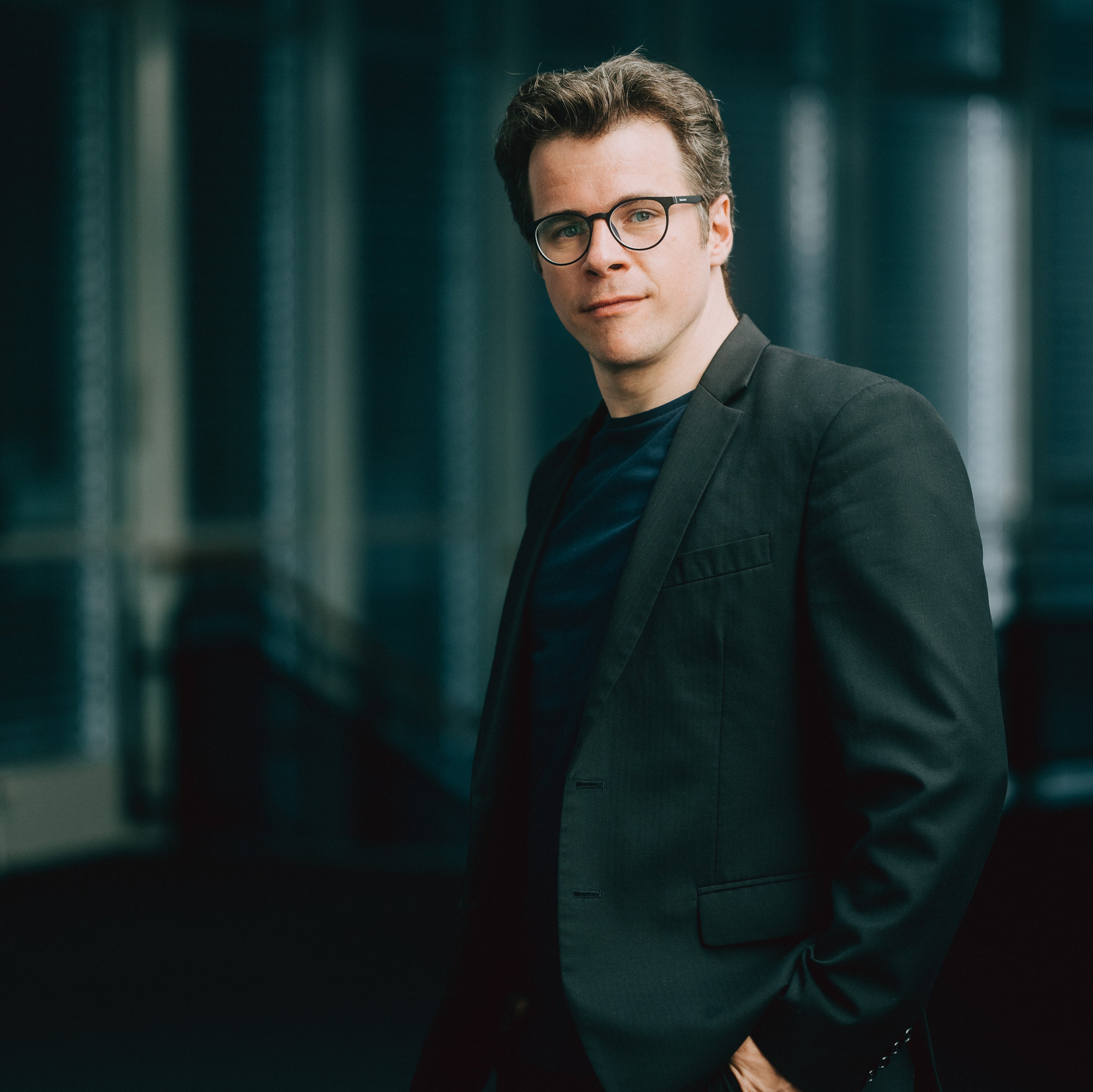 Jakub Hrůša mit Brille und dunklem Anzug blickt mit einem leichten Lächeln in die Kamera. Er steht in einer Innenumgebung mit sanftem Licht und unscharfem Hintergrund.