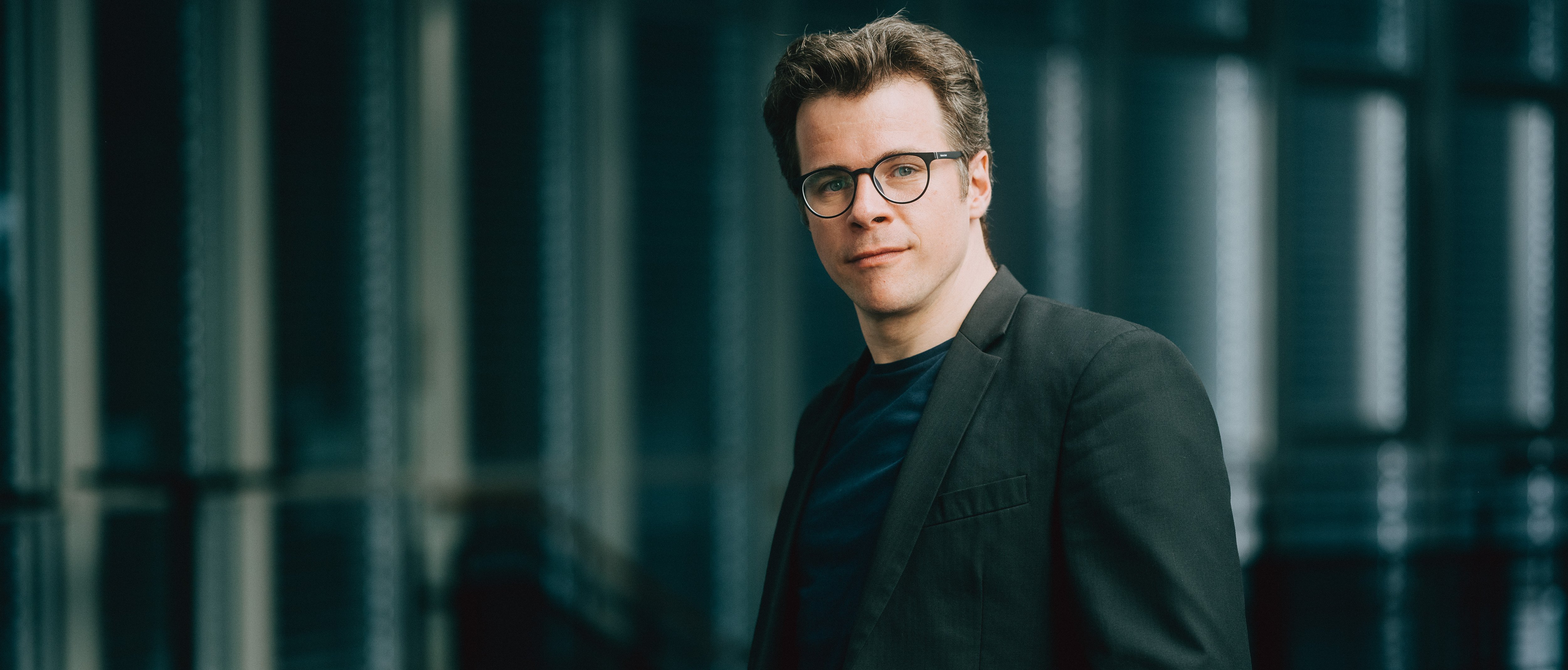 Jakub Hrůša mit Brille und dunklem Anzug blickt mit einem leichten Lächeln in die Kamera. Er steht in einer Innenumgebung mit sanftem Licht und unscharfem Hintergrund.