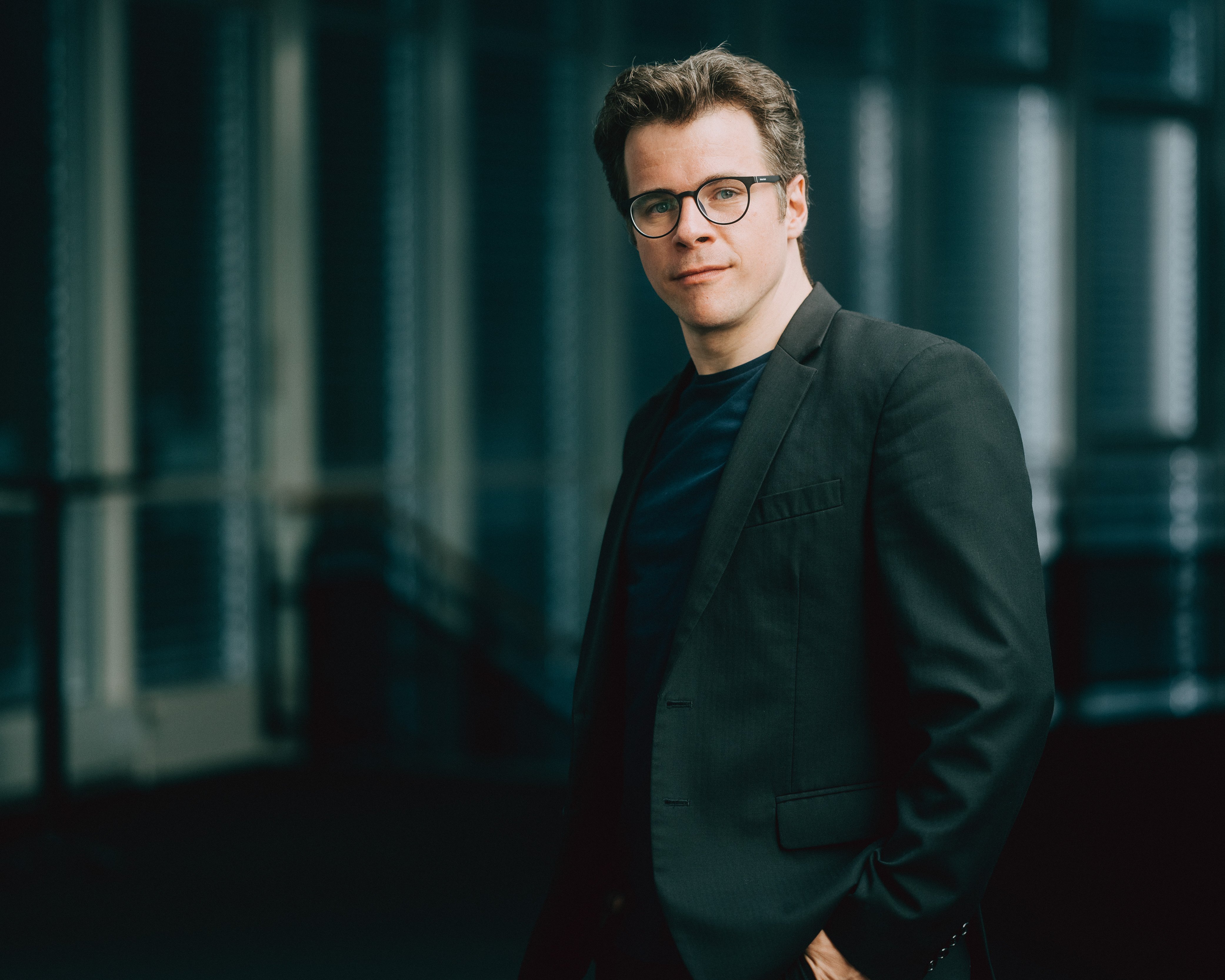 Jakub Hrůša mit Brille und dunklem Anzug blickt mit einem leichten Lächeln in die Kamera. Er steht in einer Innenumgebung mit sanftem Licht und unscharfem Hintergrund.