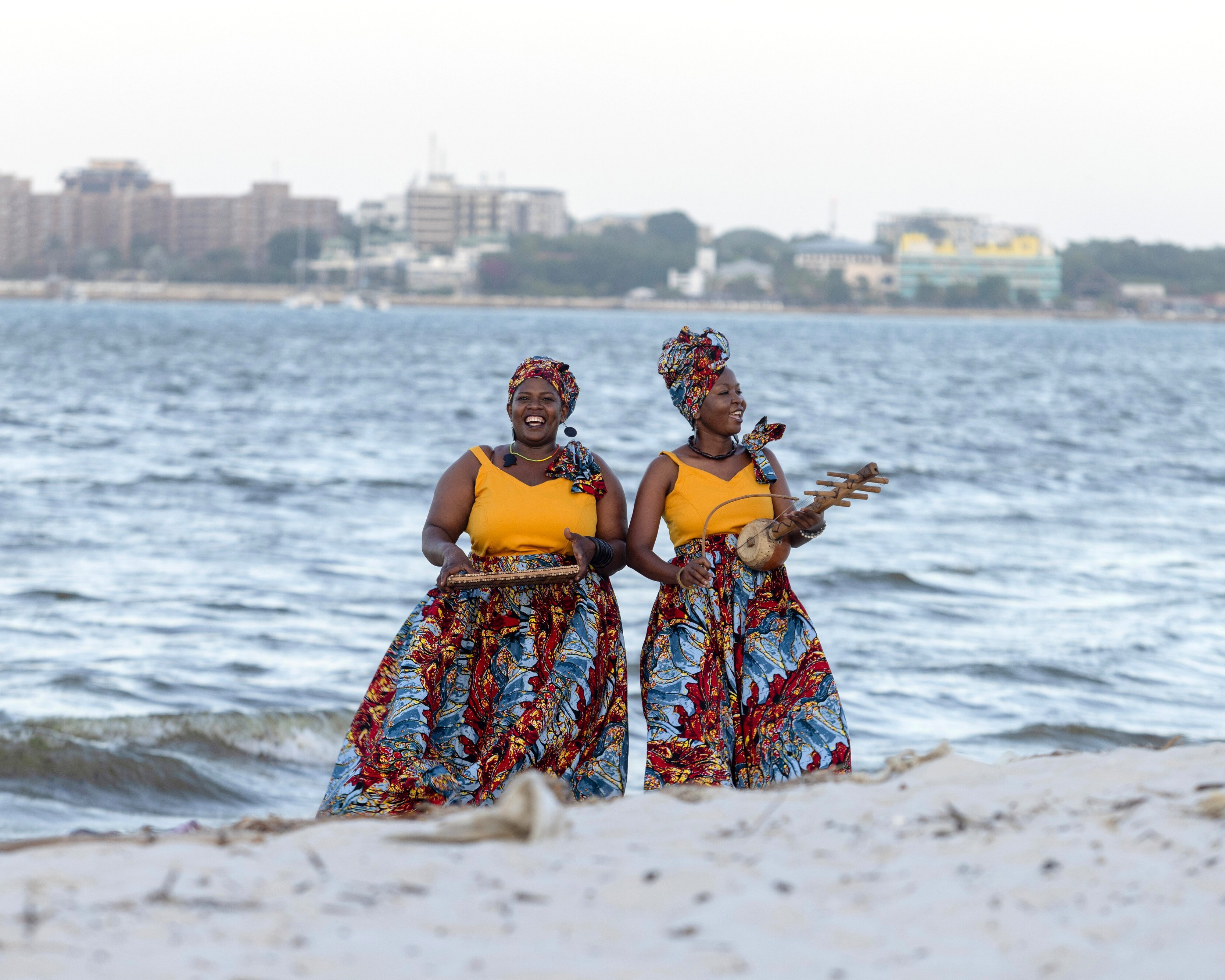 Zwei Frauen in leuchtenden, bunten Röcken und Kopftüchern stehen an einem Sandstrand in der Nähe des Ozeans und halten Tabletts mit Essen in der Hand, während im Hintergrund auf der anderen Seite des Wassers eine Stadt zu sehen ist.