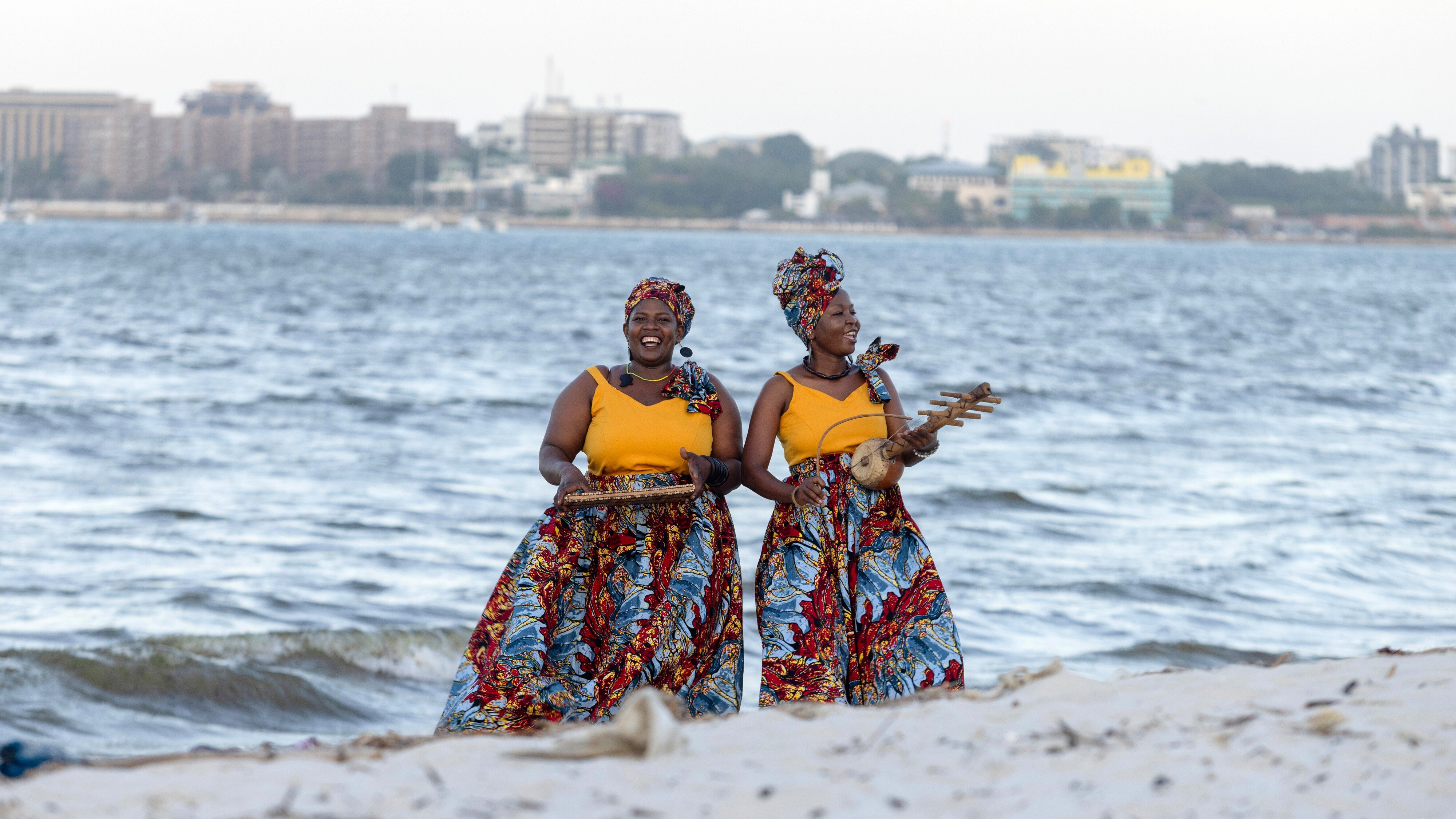 Zwei Frauen in leuchtenden, bunten Röcken und Kopftüchern stehen an einem Sandstrand in der Nähe des Ozeans und halten Tabletts mit Essen in der Hand, während im Hintergrund auf der anderen Seite des Wassers eine Stadt zu sehen ist.