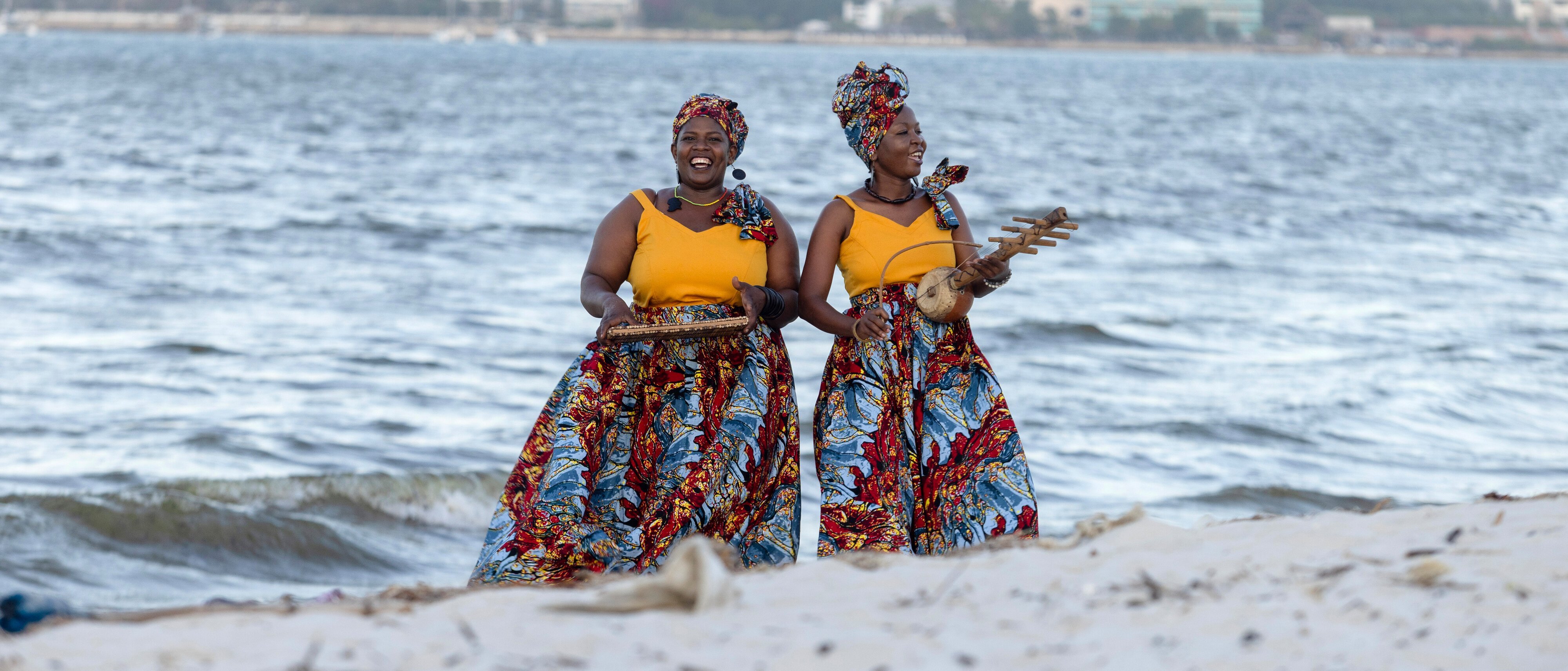Zwei Frauen in leuchtenden, bunten Röcken und Kopftüchern stehen an einem Sandstrand in der Nähe des Ozeans und halten Tabletts mit Essen in der Hand, während im Hintergrund auf der anderen Seite des Wassers eine Stadt zu sehen ist.