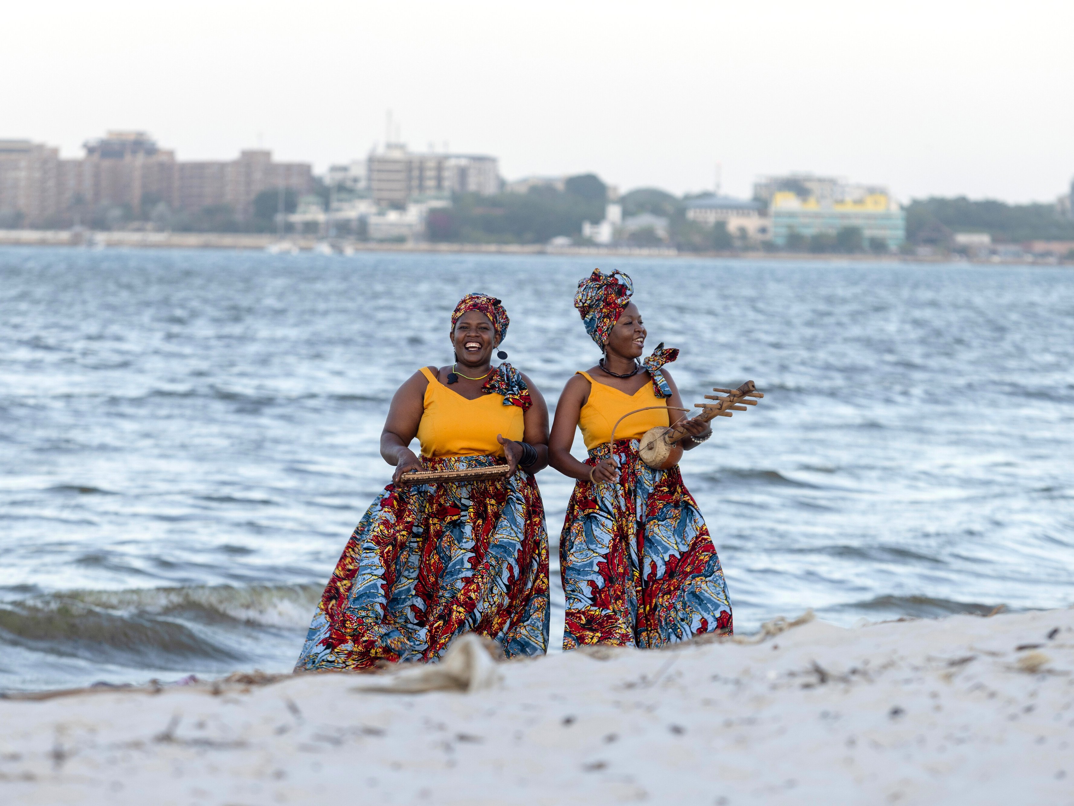 Zwei Frauen in leuchtenden, bunten Röcken und Kopftüchern stehen an einem Sandstrand in der Nähe des Ozeans und halten Tabletts mit Essen in der Hand, während im Hintergrund auf der anderen Seite des Wassers eine Stadt zu sehen ist.