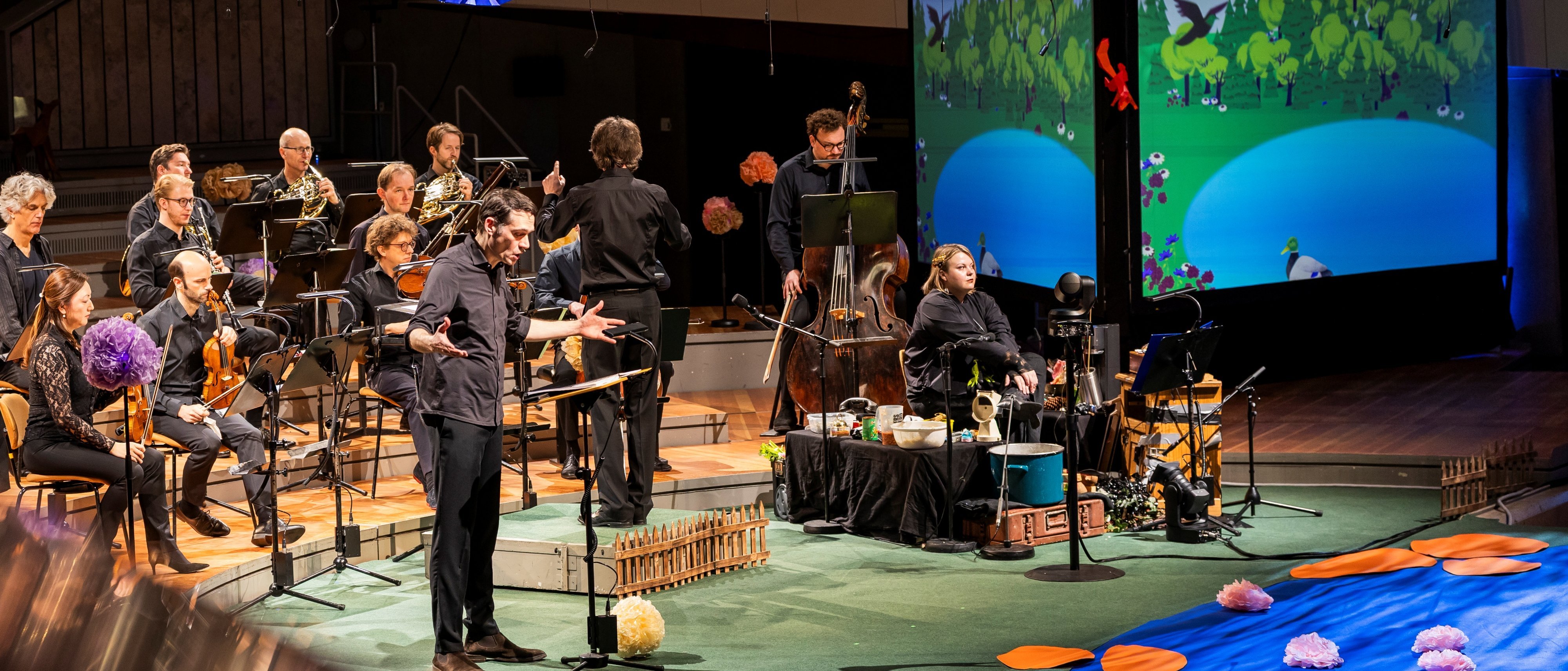 Ein Kammerorchester spielt auf der Bühne zusammen mit einem Sprecher, einer Geräuschemacherin,  Requisiten und einem Bildschirm, auf dem eine animierte Szenerie eines Teiches mit Vögeln und Bäumen zu sehen ist. Die Bühne ist mit orangefarbenen Seerosenblättern und lila Blumen geschmückt.
