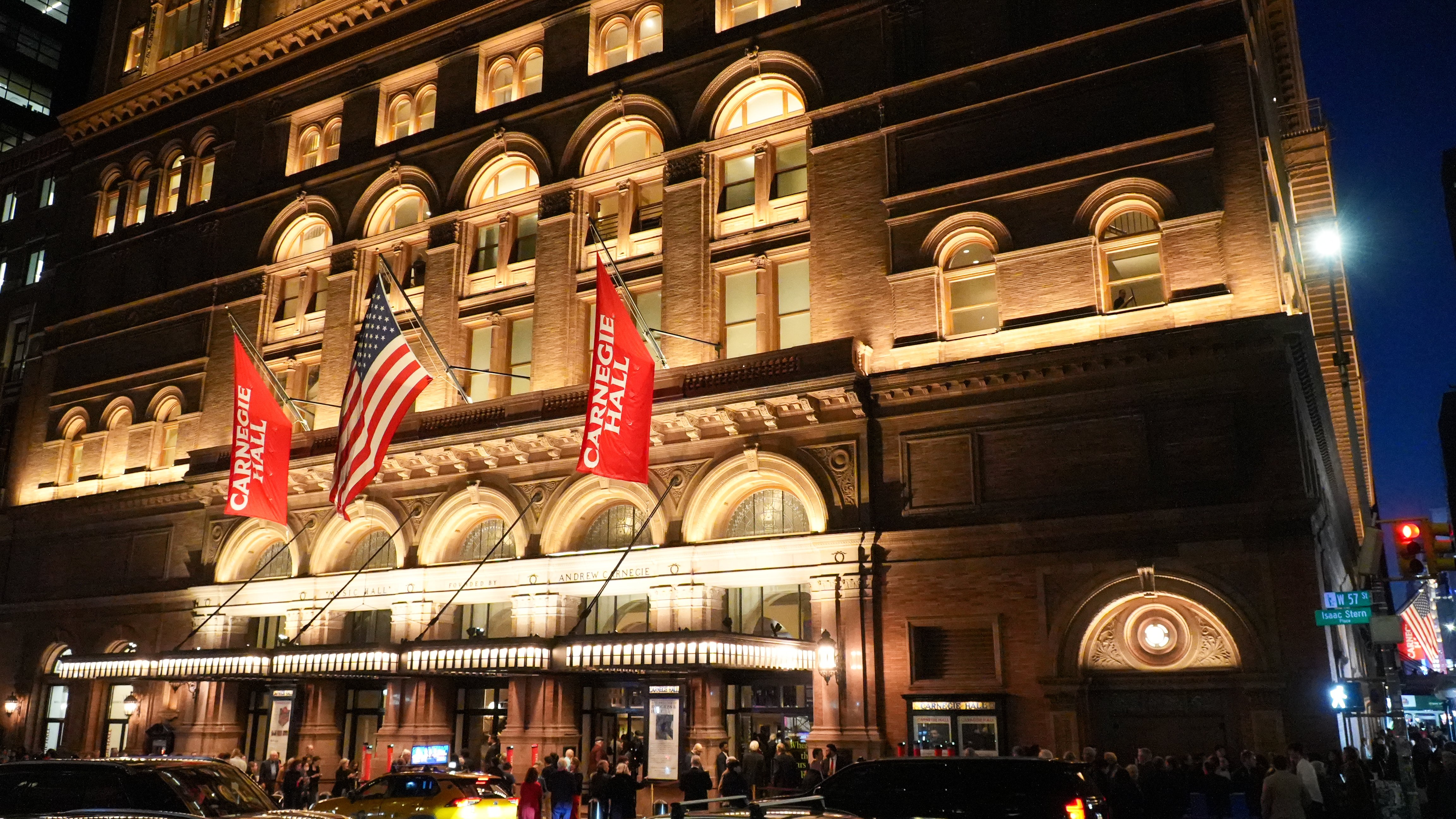 Fassade der Carnegie Hall bei Nacht, hell erleuchtet. Im Vordergrund sind Autos und Fußgänger zu sehen.