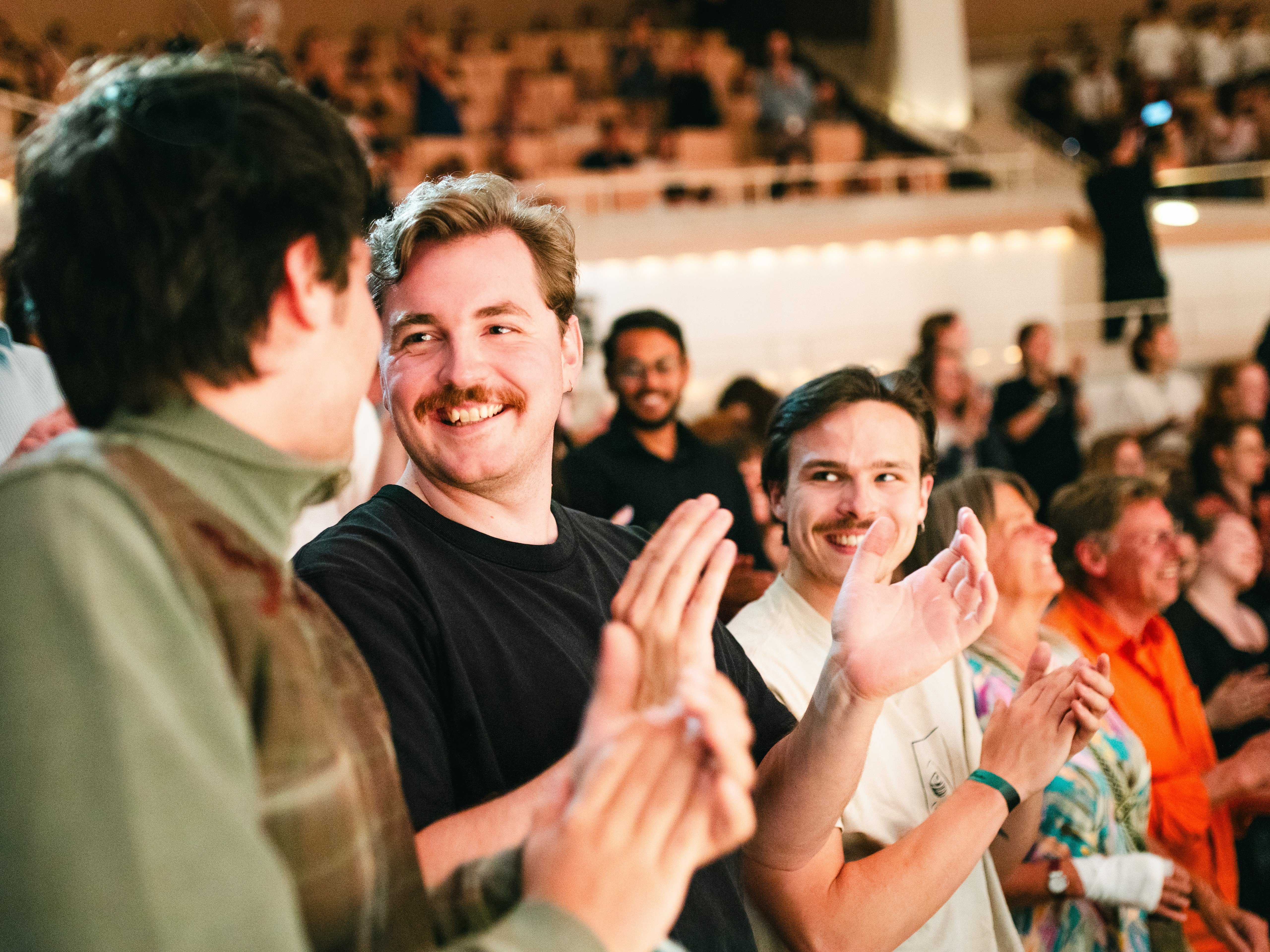 A group of people are standing and smiling as they applaud at an indoor event. The background shows a crowd sitting in an auditorium.