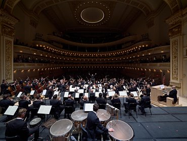 Ein Orchester spielt auf der Bühne eines prächtigen, mehrstöckigen Konzertsaals; die Aufnahme zeigt den Blick von hinter der Schlagzeuggruppe.