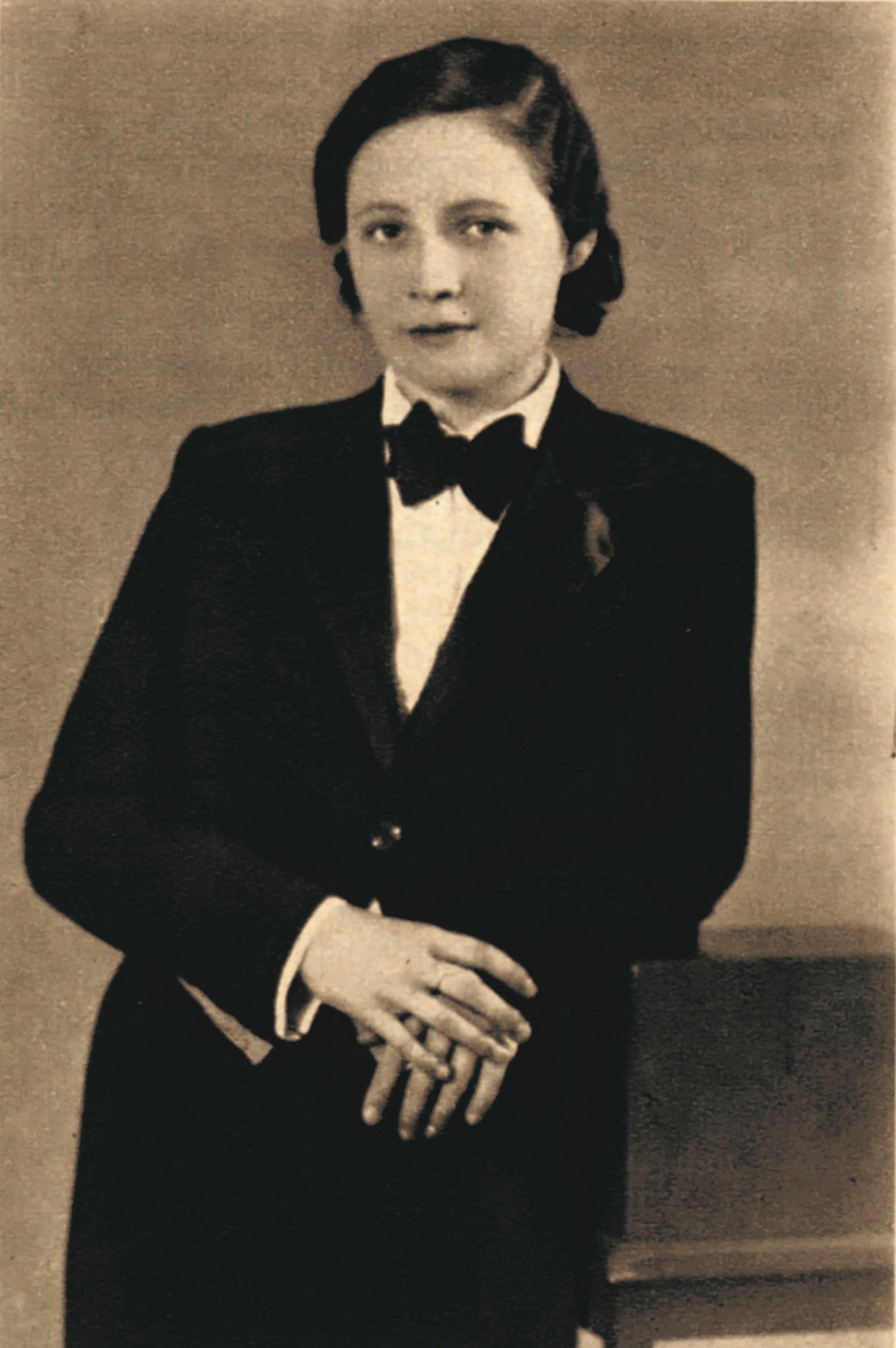A woman with short dark hair, wearing a tuxedo and bow tie, stands with hands folded in front of a neutral background in a sepia-toned, vintage-style portrait.