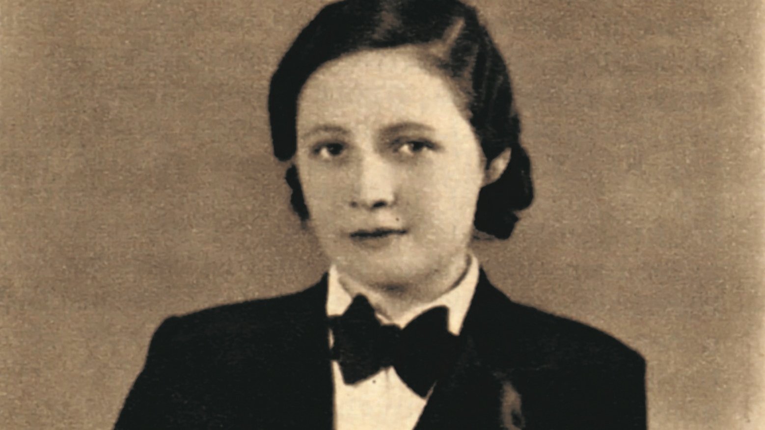 A woman with short dark hair, wearing a tuxedo and bow tie, stands with hands folded in front of a neutral background in a sepia-toned, vintage-style portrait.