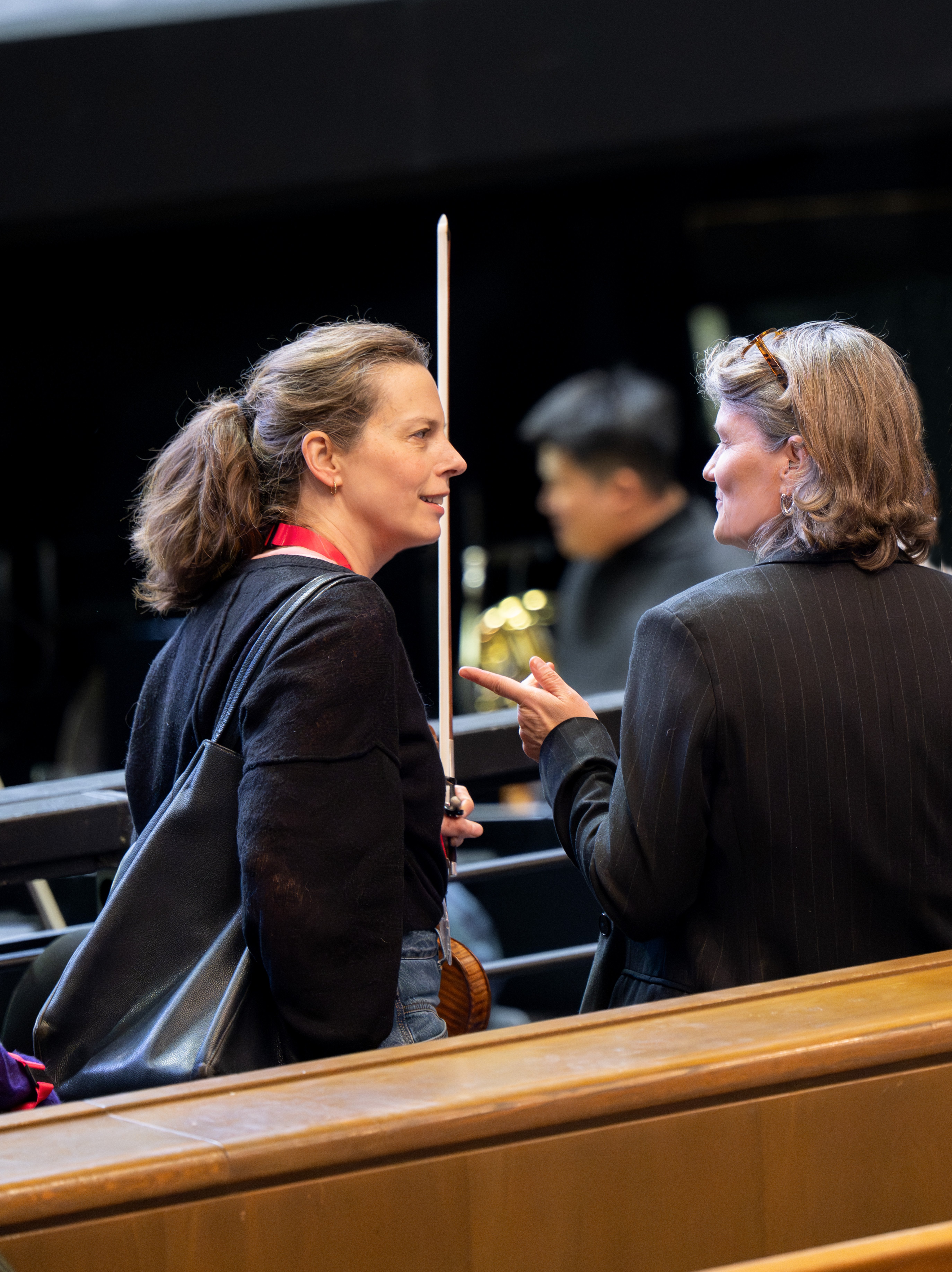 Zwei Frauen unterhalten sich im Saal. Die eine hält eine Geige und einen Bogen in der Hand und trägt eine Tasche auf der Schulter. 