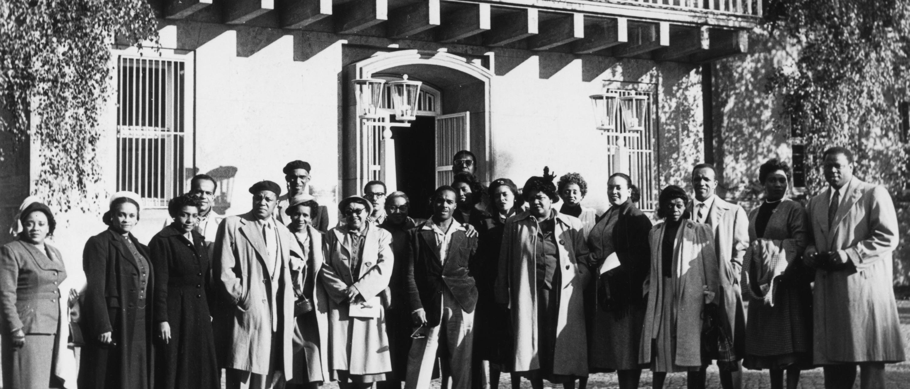 A group of Black men and women in coats pose together outside a large building with balconies and hanging lanterns; vines cover part of the building’s façade.