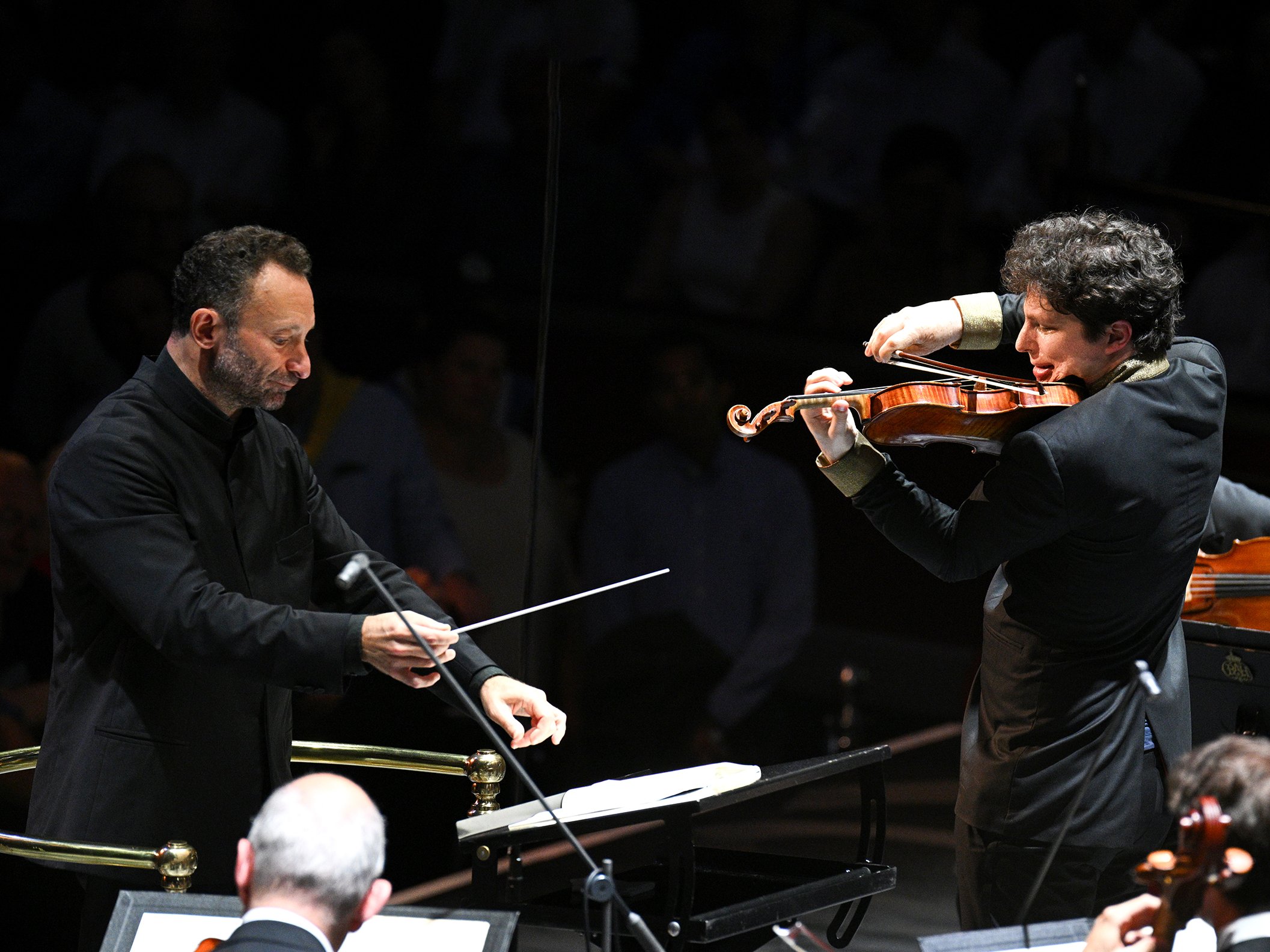 A conductor leads an orchestra while a violinist performs a solo. Both are wearing black suits, and the background shows an audience in dim lighting.