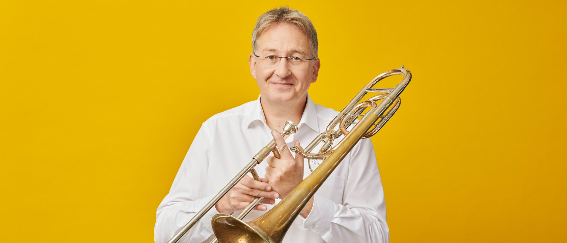 Ein Mann mit blondem Haar in einem weißen Hemd hält eine Posaune vor einem leuchtend gelben Hintergrund.