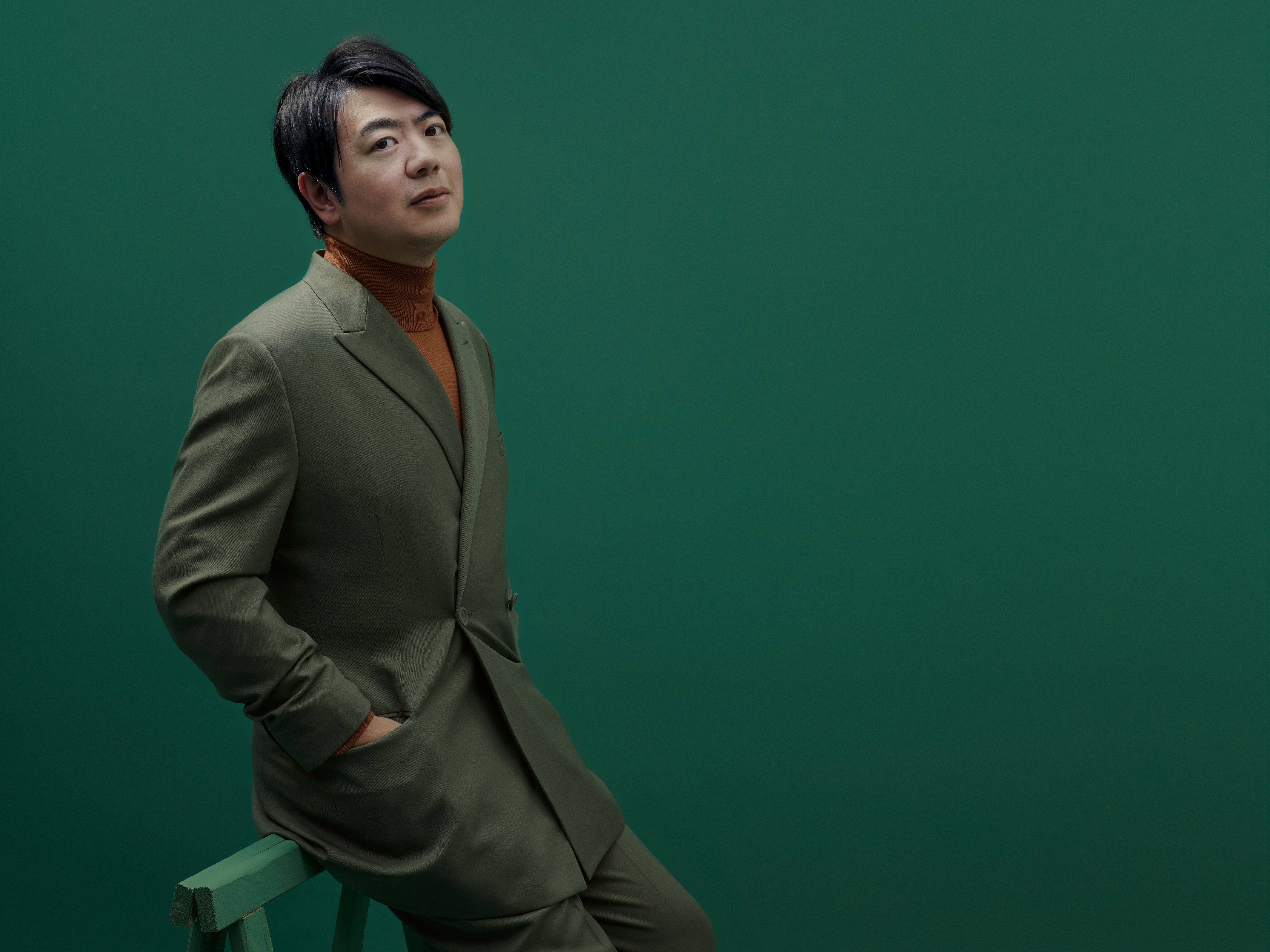Lang Lang in einem grünen Anzug und einem braunen Rollkragenpullover sitzt auf einem grünen Hocker vor einem grünen Hintergrund. Er hat kurzes, dunkles Haar und sein Gesichtsausdruck ist neutral, während er leicht nach oben blickt.