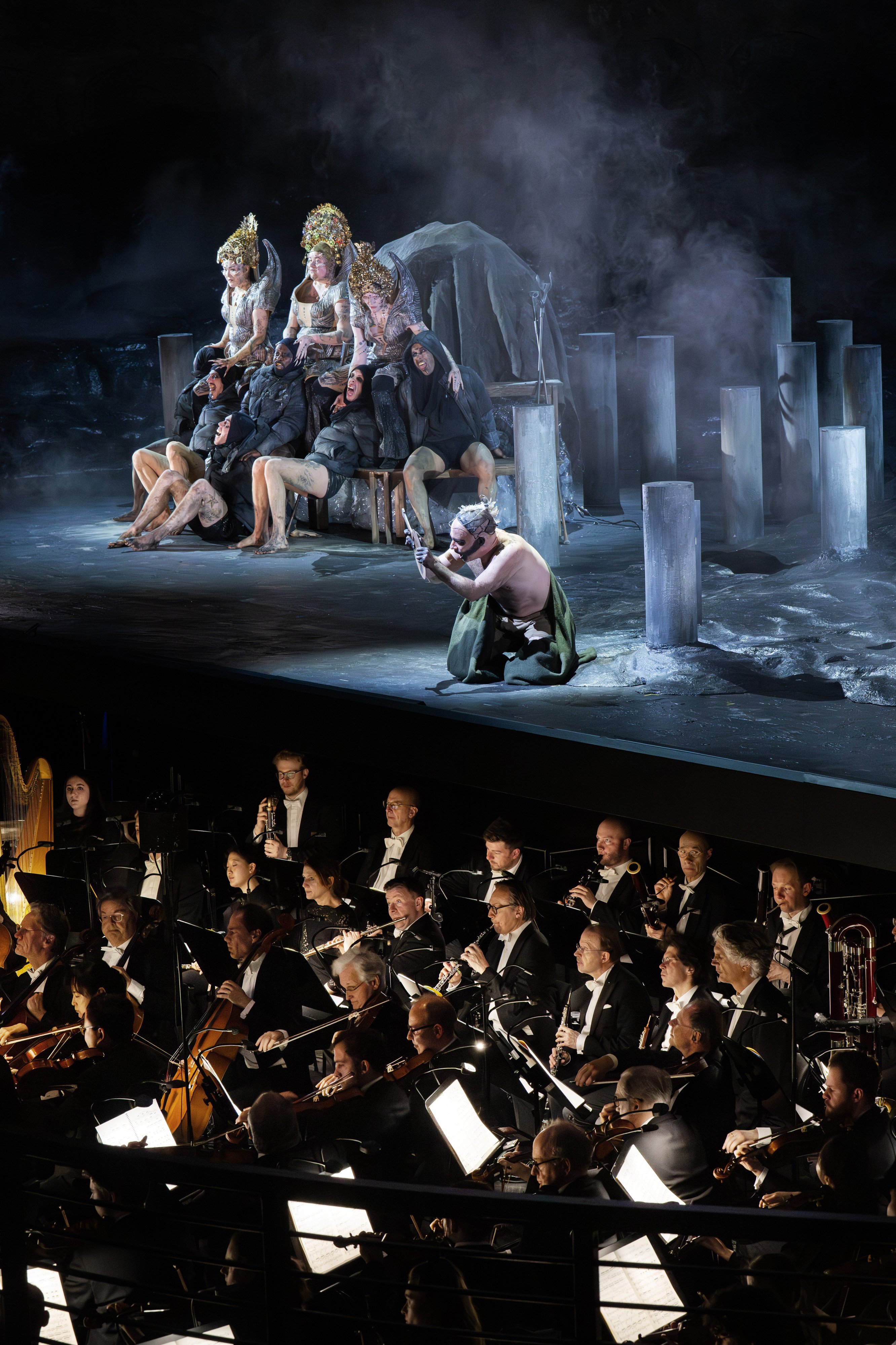 A staged opera scene shows costumed performers on a dramatic set with pillars and fog, above an orchestra pit where musicians in formal wear play various instruments.