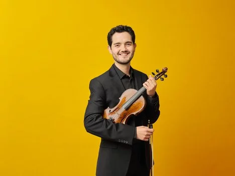 A smiling man in a black suit holds a violin and bow against a bright yellow background.
