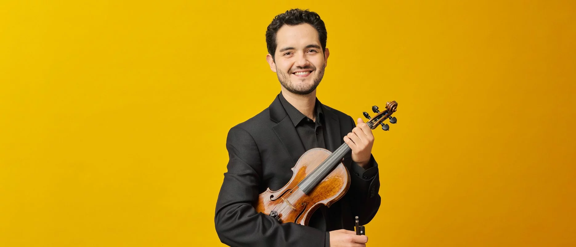 A smiling man in a black suit holds a violin and bow against a bright yellow background.