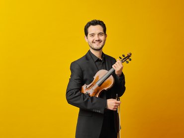 A smiling man in a black suit holds a violin and bow against a bright yellow background.