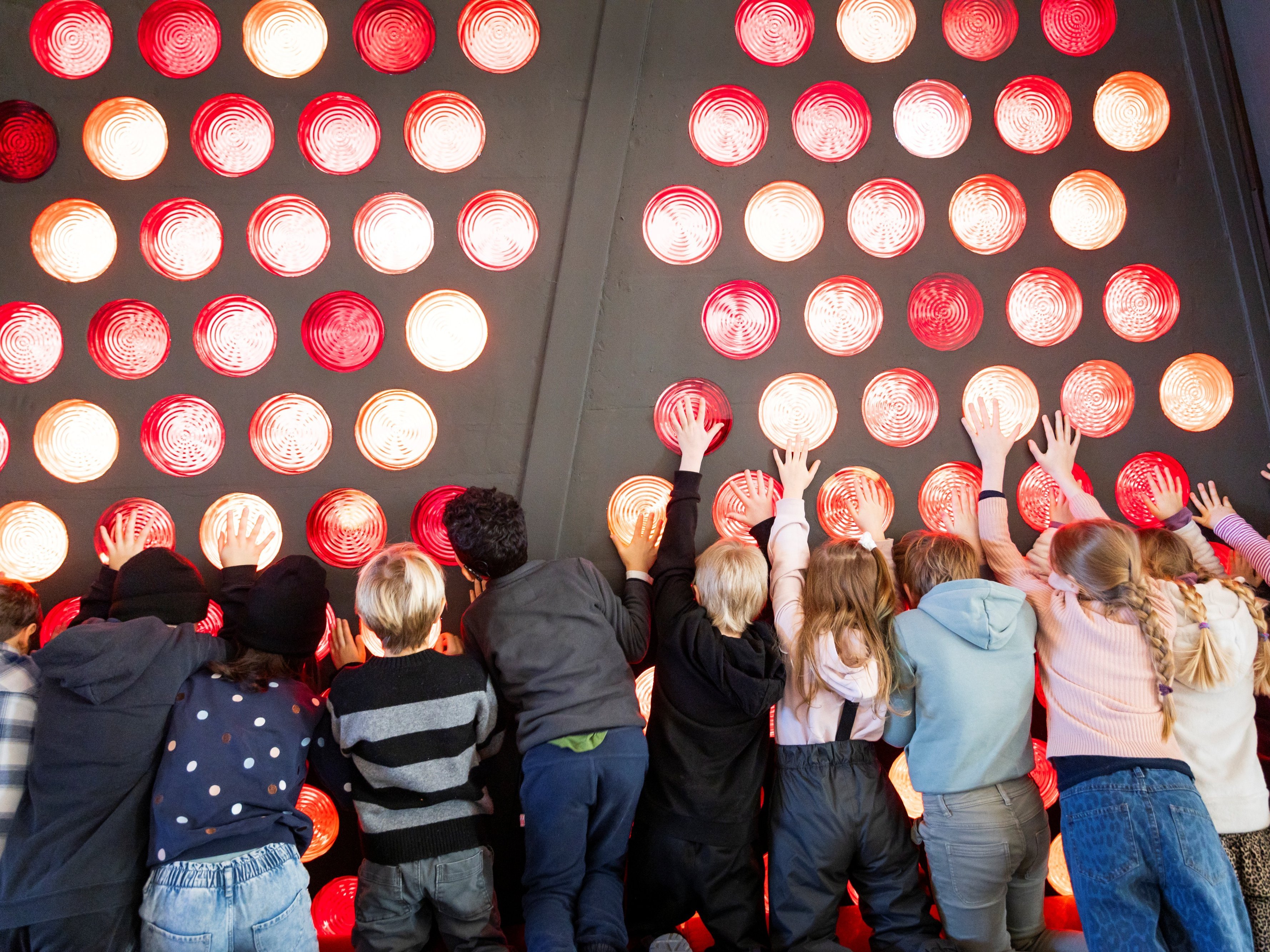Eine Gruppe von Kindern steht in einer Reihe und streckt ihre Hände in Richtung einer großen Wand, die mit leuchtenden roten und orangefarbenen Glasbausteinen bedeckt ist. Die Kinder scheinen spielerisch mit der Wand zu interagieren.