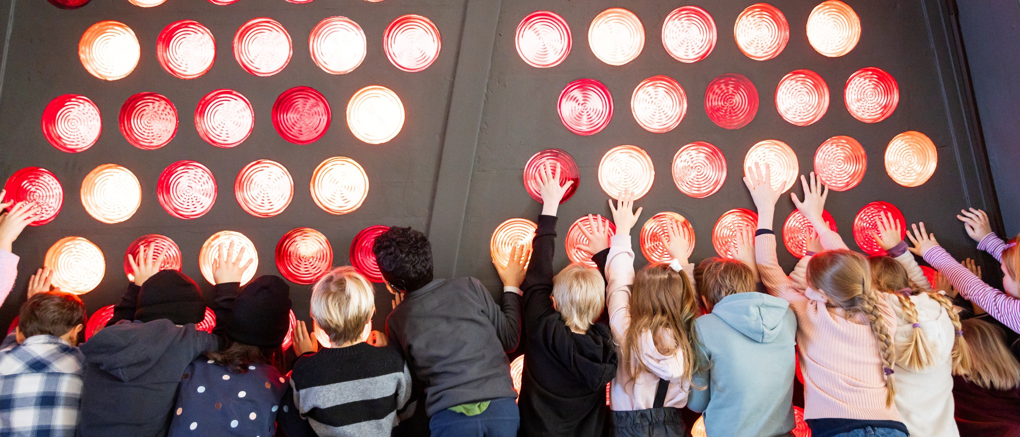 Eine Gruppe von Kindern steht in einer Reihe und streckt ihre Hände in Richtung einer großen Wand, die mit leuchtenden roten und orangefarbenen Glasbausteinen bedeckt ist. Die Kinder scheinen spielerisch mit der Wand zu interagieren.
