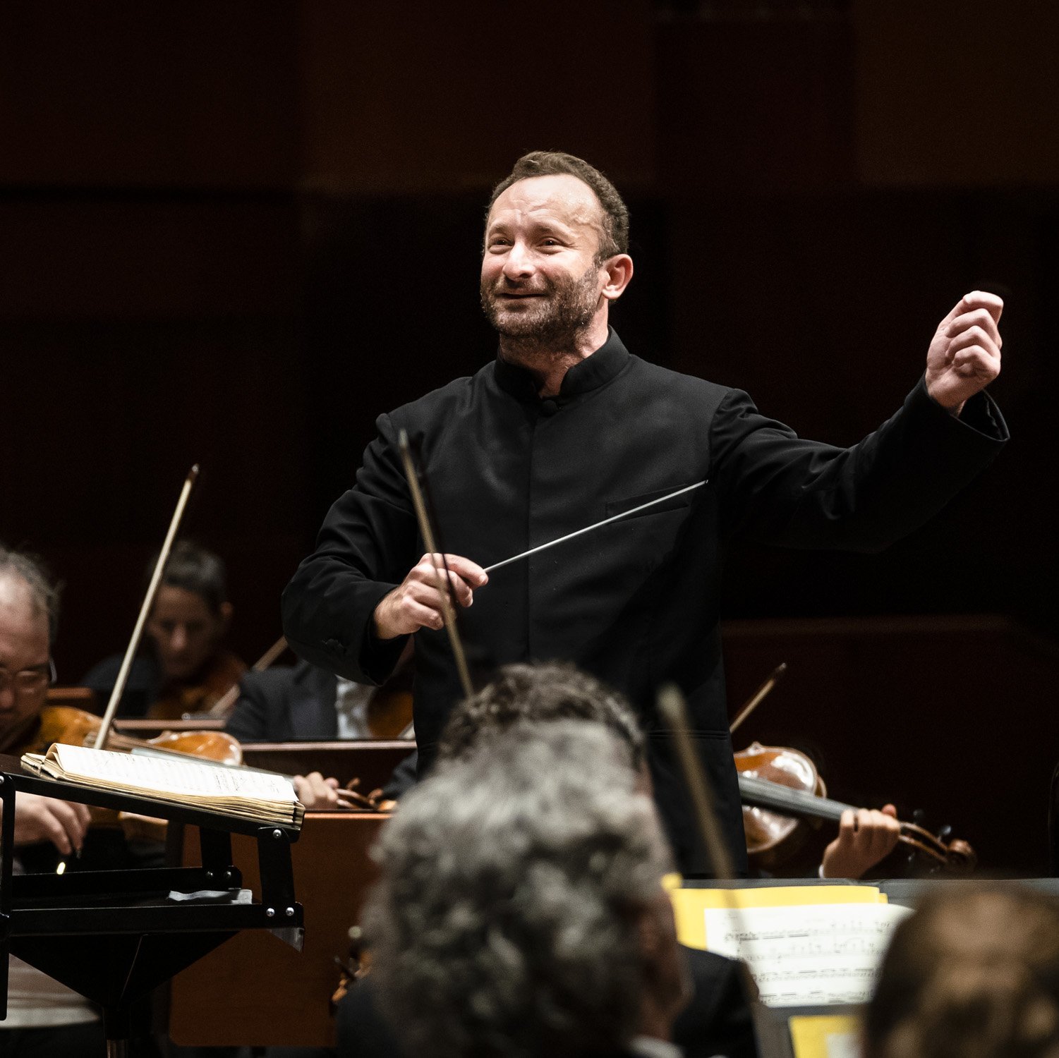 Kirill Petrenko energetically conducts the orchestra. He holds a baton and gestures with his other hand, surrounded by musicians playing various string instruments.
