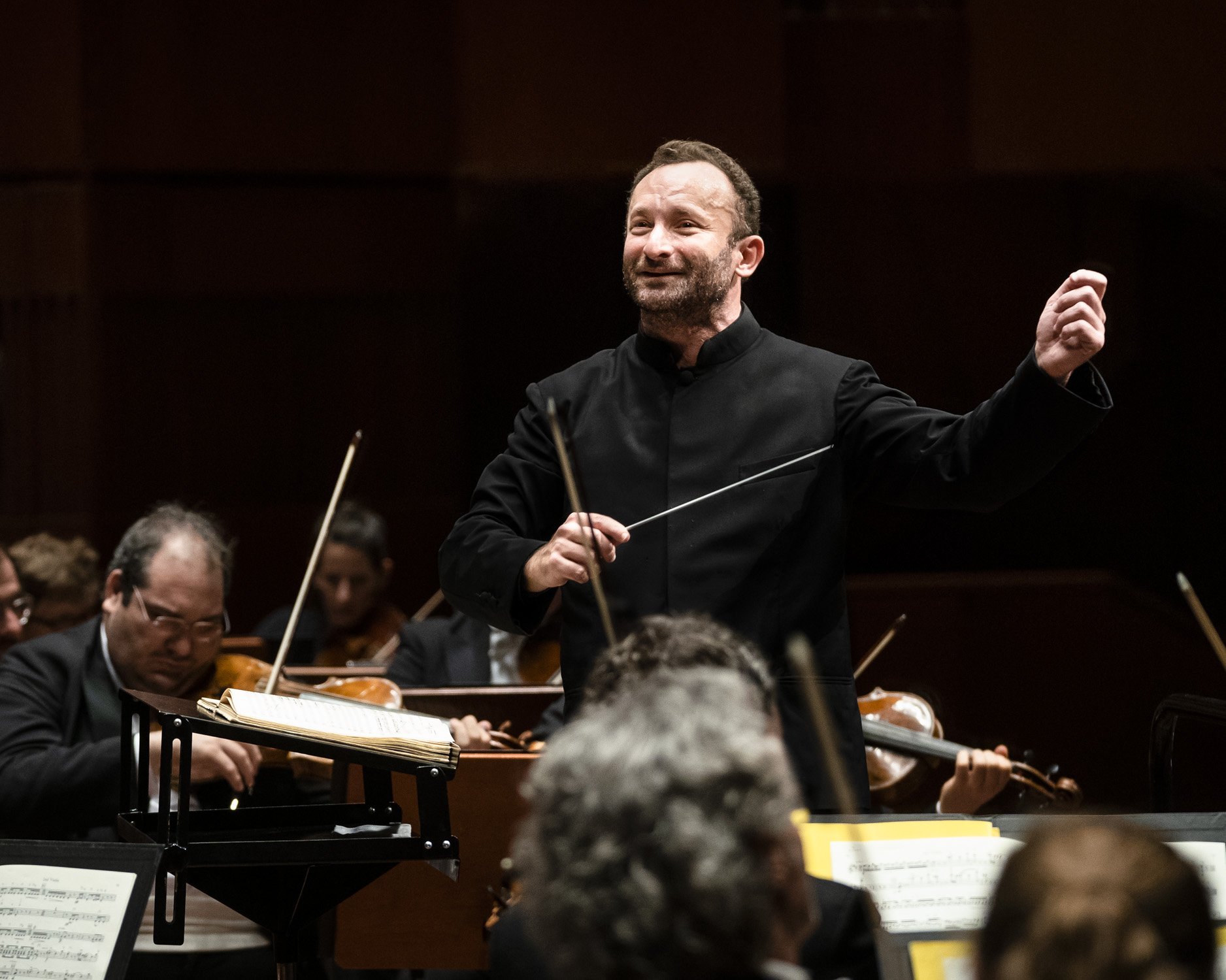 Kirill Petrenko leitet energisch das Orchester. Er hält einen Taktstock und gestikuliert mit der anderen Hand, umgeben von Musikern, die verschiedene Streichinstrumente spielen. 