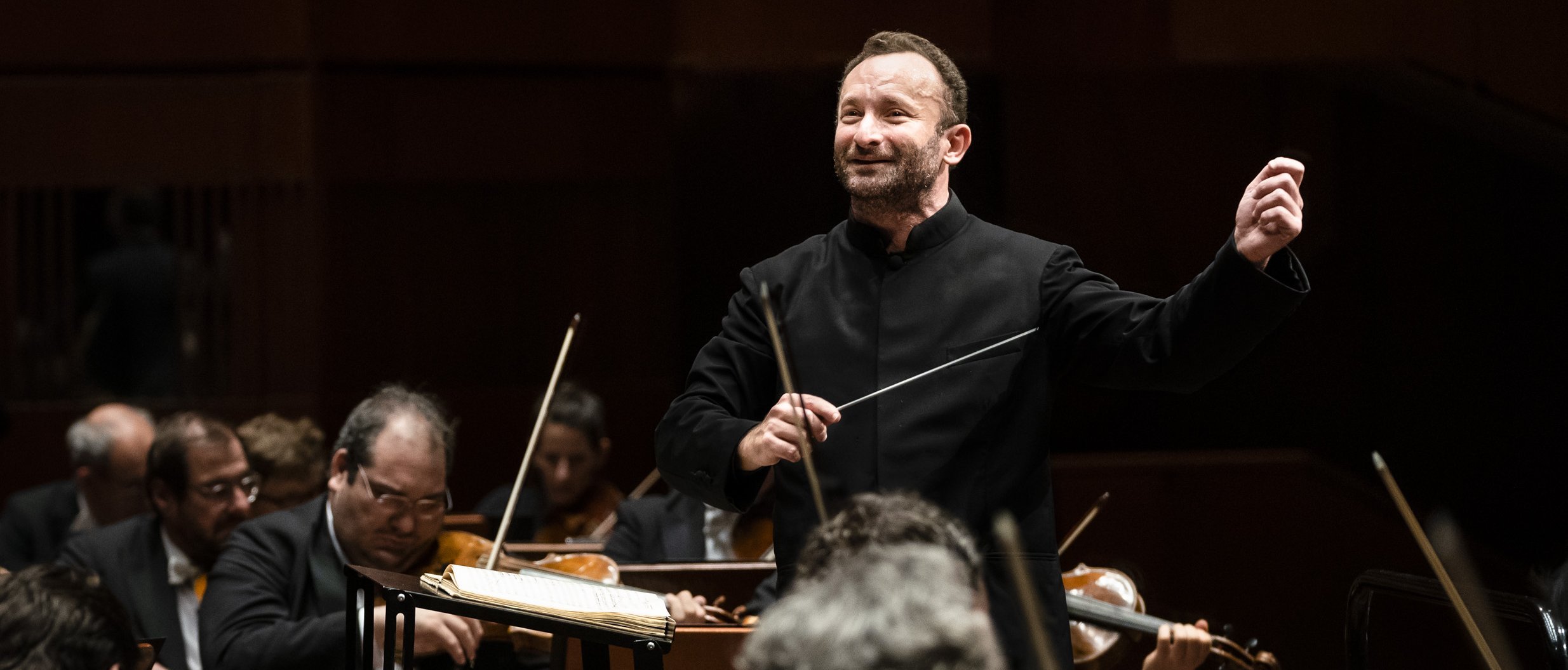 Kirill Petrenko leitet energisch das Orchester. Er hält einen Taktstock und gestikuliert mit der anderen Hand, umgeben von Musikern, die verschiedene Streichinstrumente spielen. 