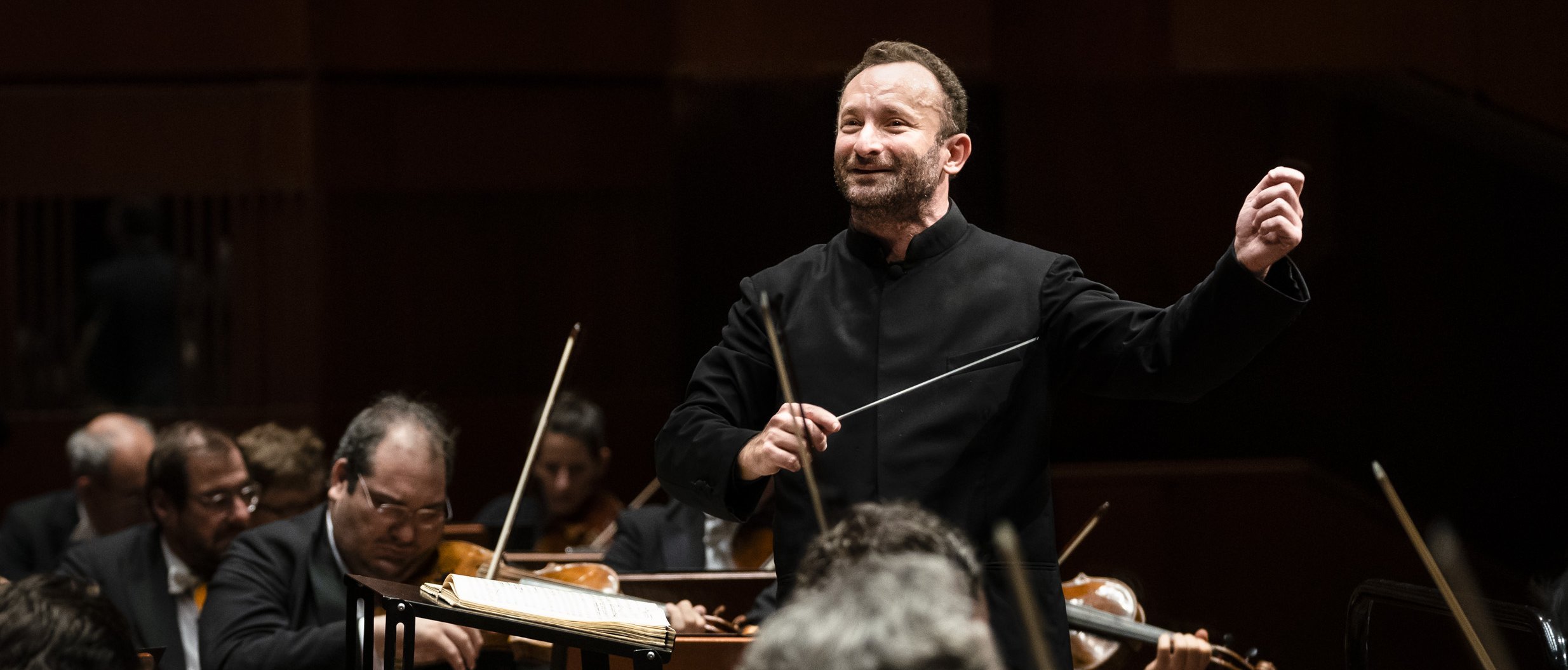 Kirill Petrenko leitet energisch das Orchester. Er hält einen Taktstock und gestikuliert mit der anderen Hand, umgeben von Musikern, die verschiedene Streichinstrumente spielen. 
