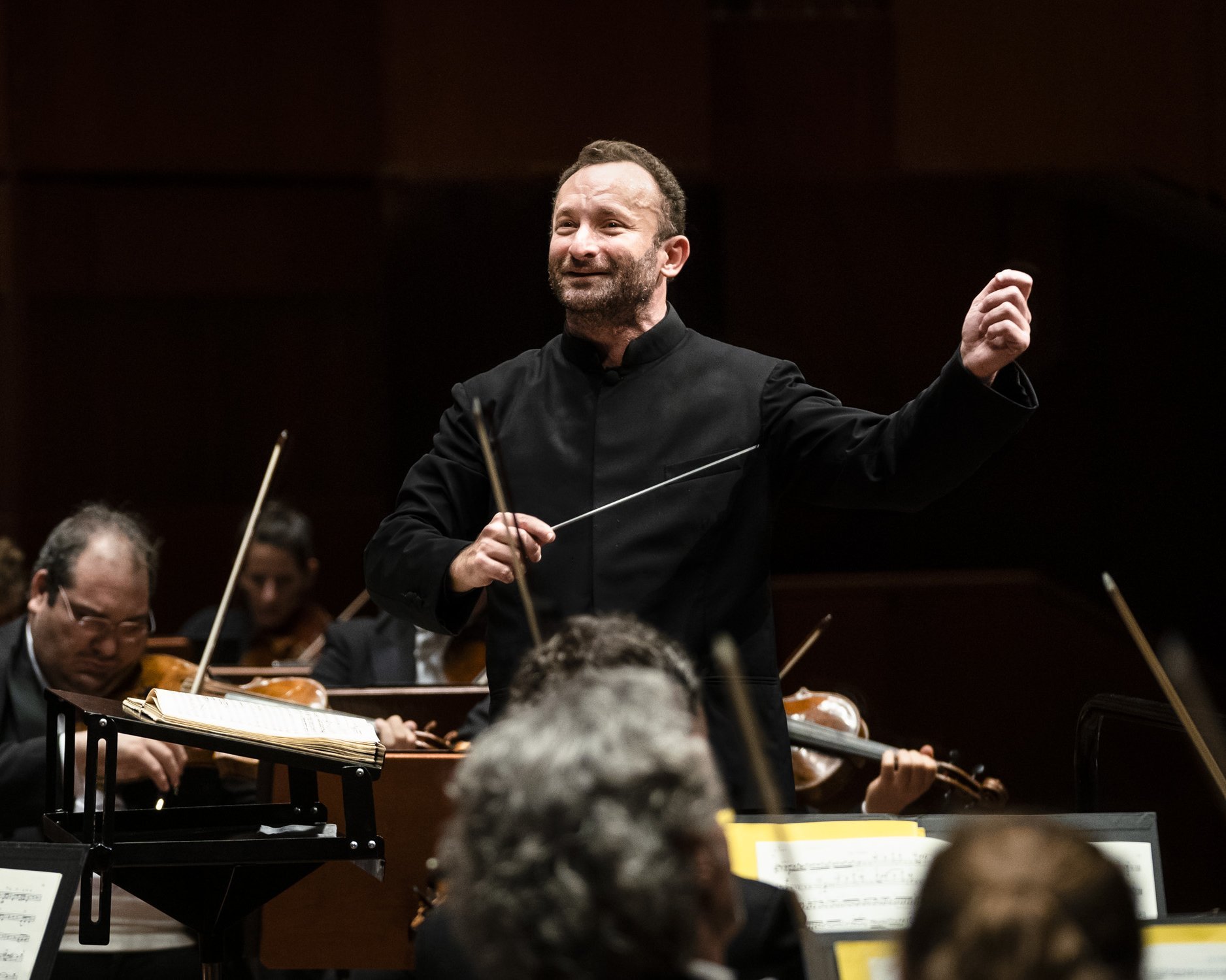 Kirill Petrenko leitet energisch das Orchester. Er hält einen Taktstock und gestikuliert mit der anderen Hand, umgeben von Musikern, die verschiedene Streichinstrumente spielen. 