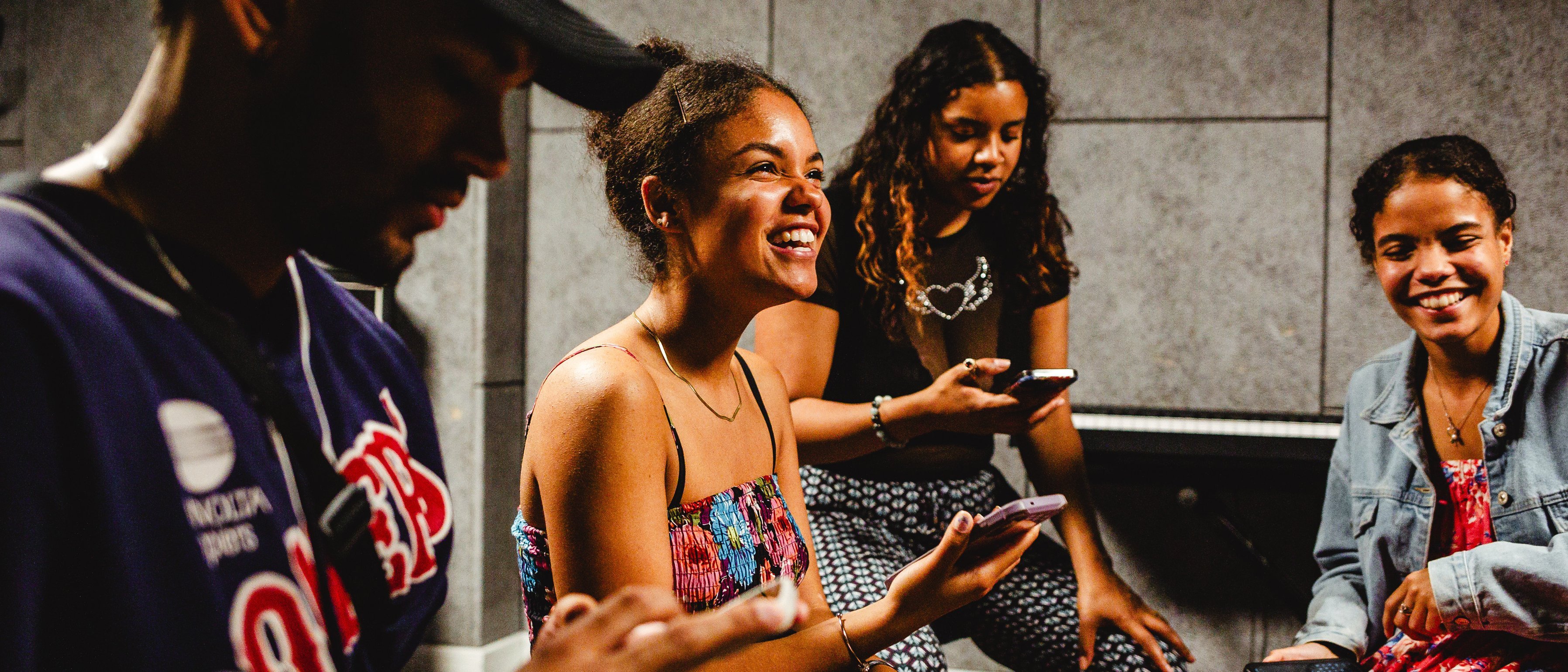 The picture shows four young people sitting together and using their mobile phones. Two of them are laughing and appear cheerful, while the others are looking at their smartphones. They are in a room with grey walls.