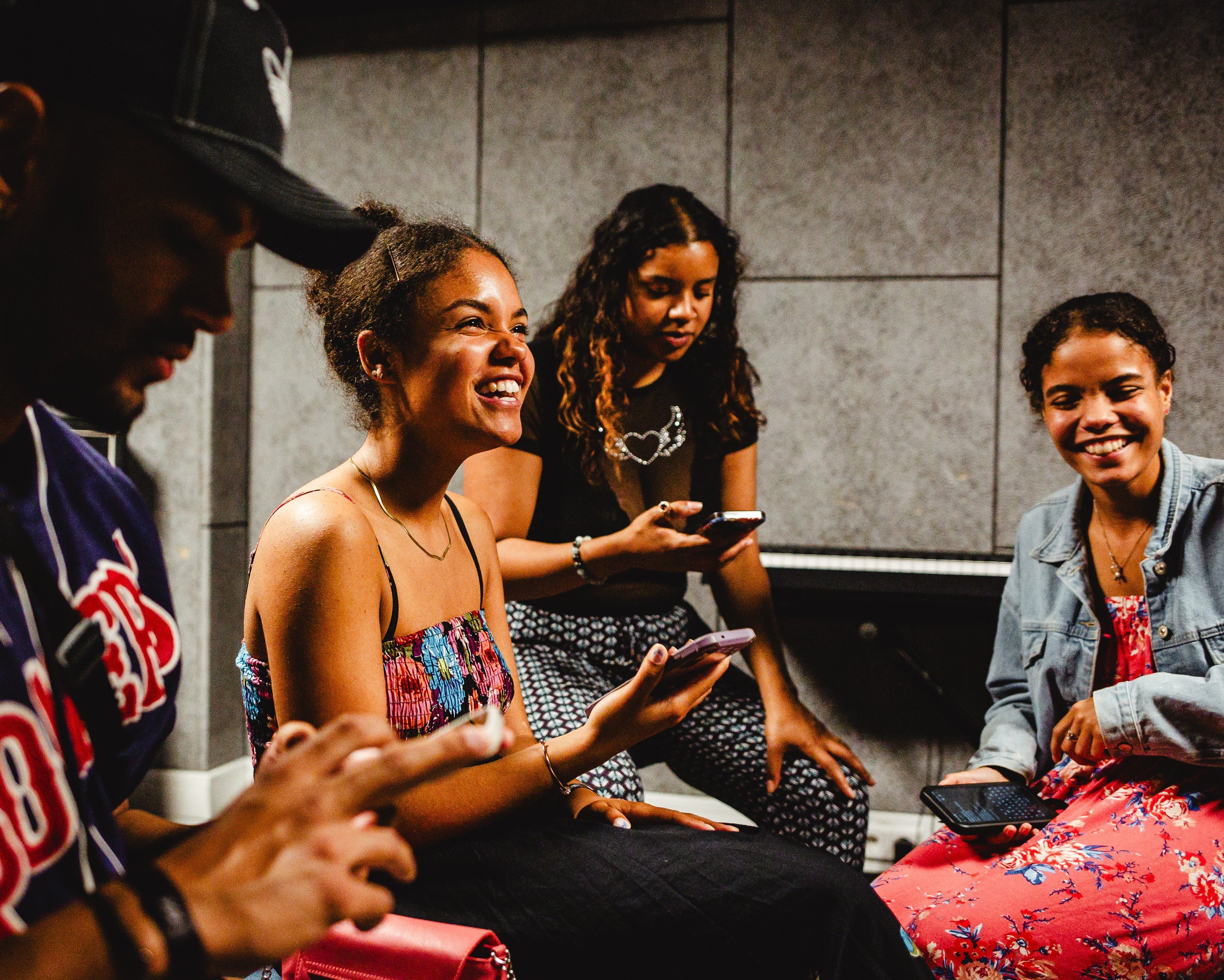 Auf dem Bild sieht man vier junge Menschen, die zusammen sitzen und ihre Handys nutzen. Zwei von ihnen lachen und wirken fröhlich, während die anderen auf ihre Smartphones schauen. Sie befinden sich in einem Raum mit grauen Wänden.