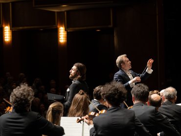 Ein Dirigent leitet ein Orchester auf der Bühne, auf der Musiker Streichinstrumente spielen und ein Publikum in einem schwach beleuchteten Konzertsaal zusieht. Ein Sänger steht mit einer Notenmappe da.
