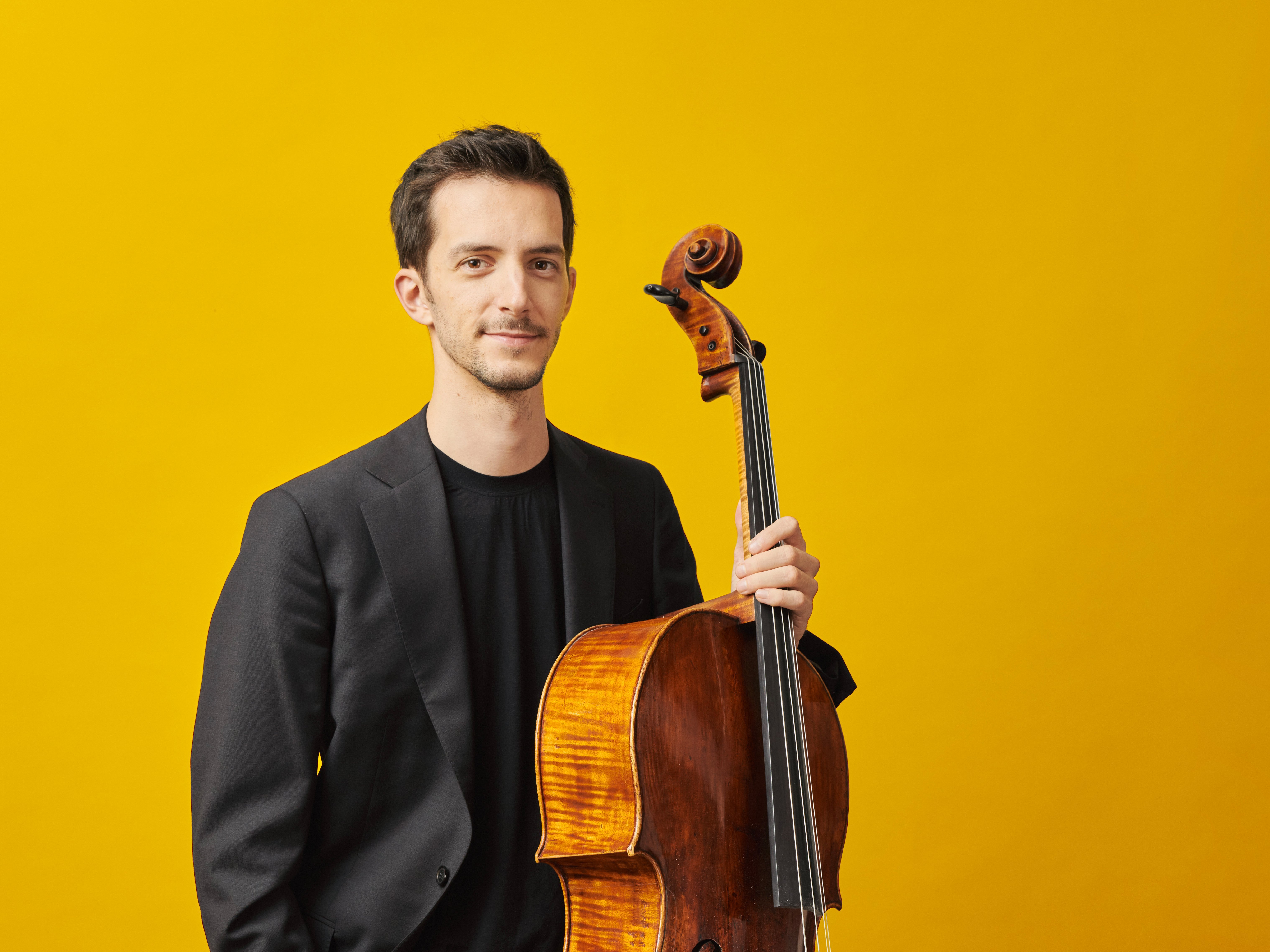 Solocellist Bruno Delepelaire in schwarzem Hemd und Jackett. Er hält sein Celle in die Höhe und steht vor einem leuchtend gelben Hintergrund. 