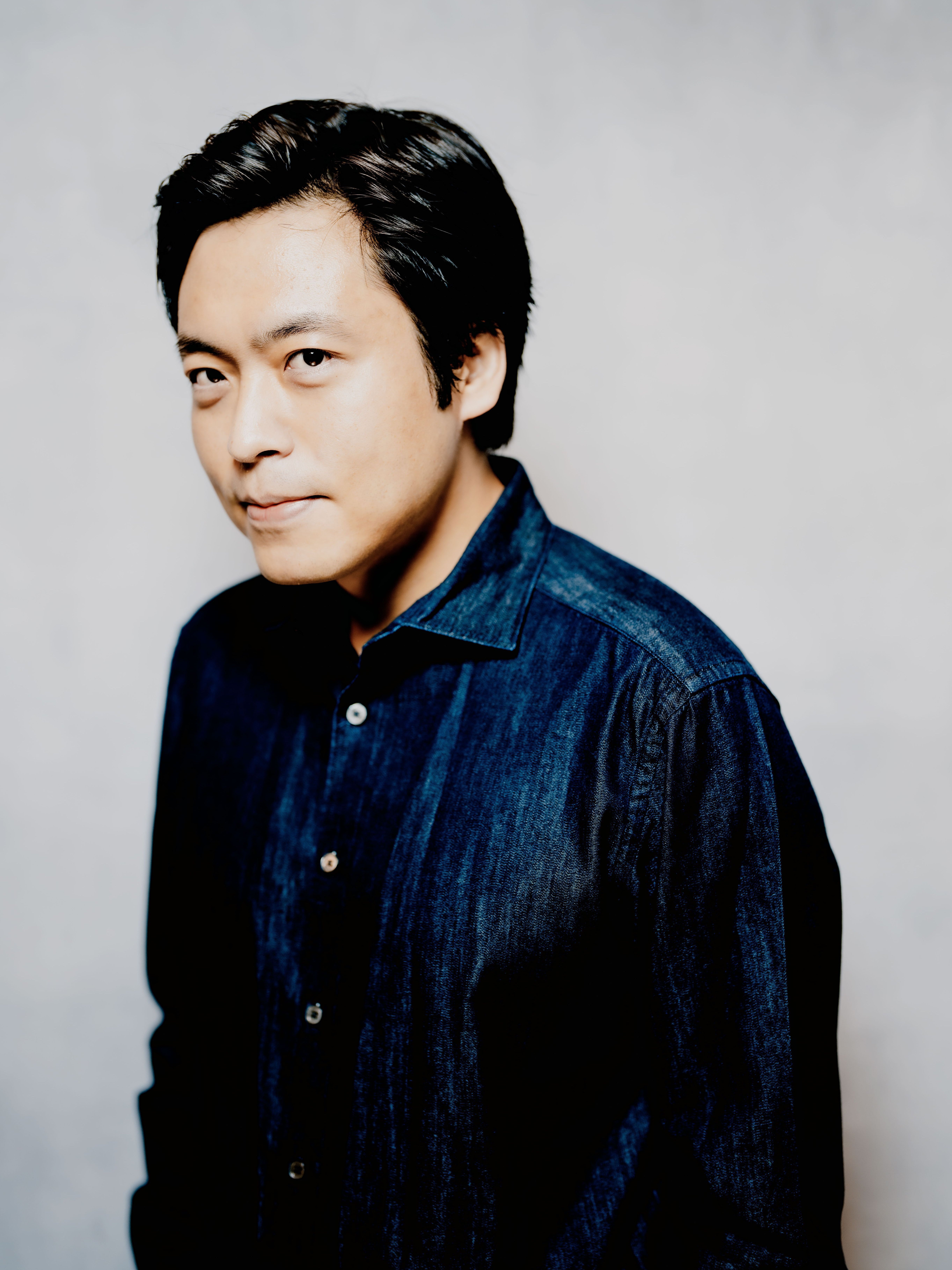 Sunwook Kim mit kurzen dunklen Haaren und einem dunkelblauen Jeanshemd blickt mit einem subtilen Lächeln in die Kamera. Der Hintergrund ist in einer hellen, neutralen Farbe gehalten.