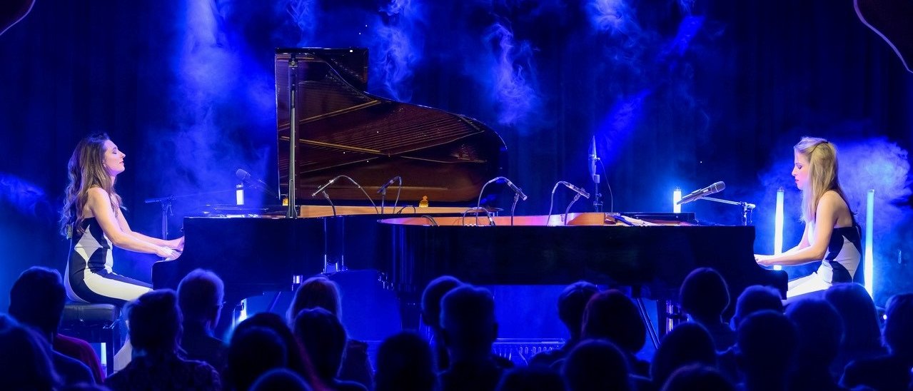 Two women play grand pianos on a stage with blue lighting and fog, facing each other. An audience watches the performance in a darkened room. Microphones are set up around the pianos.
