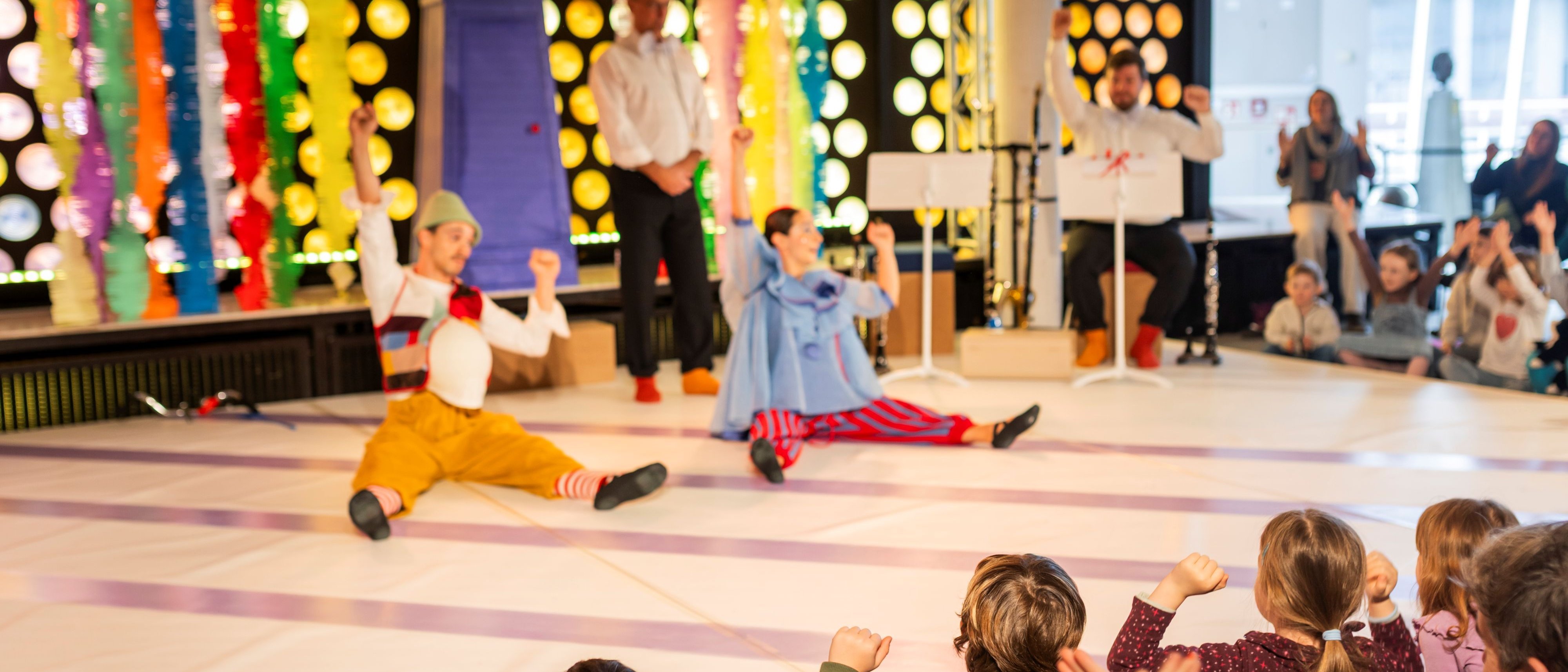 Zwei Darstellende treten auf der Bühne vor einem Publikum von Kindern auf, mit bunten Dekorationen und hellen Lichtern im Hintergrund. Die Kinder schauen gespannt zu, einige heben aufgeregt die Arme.