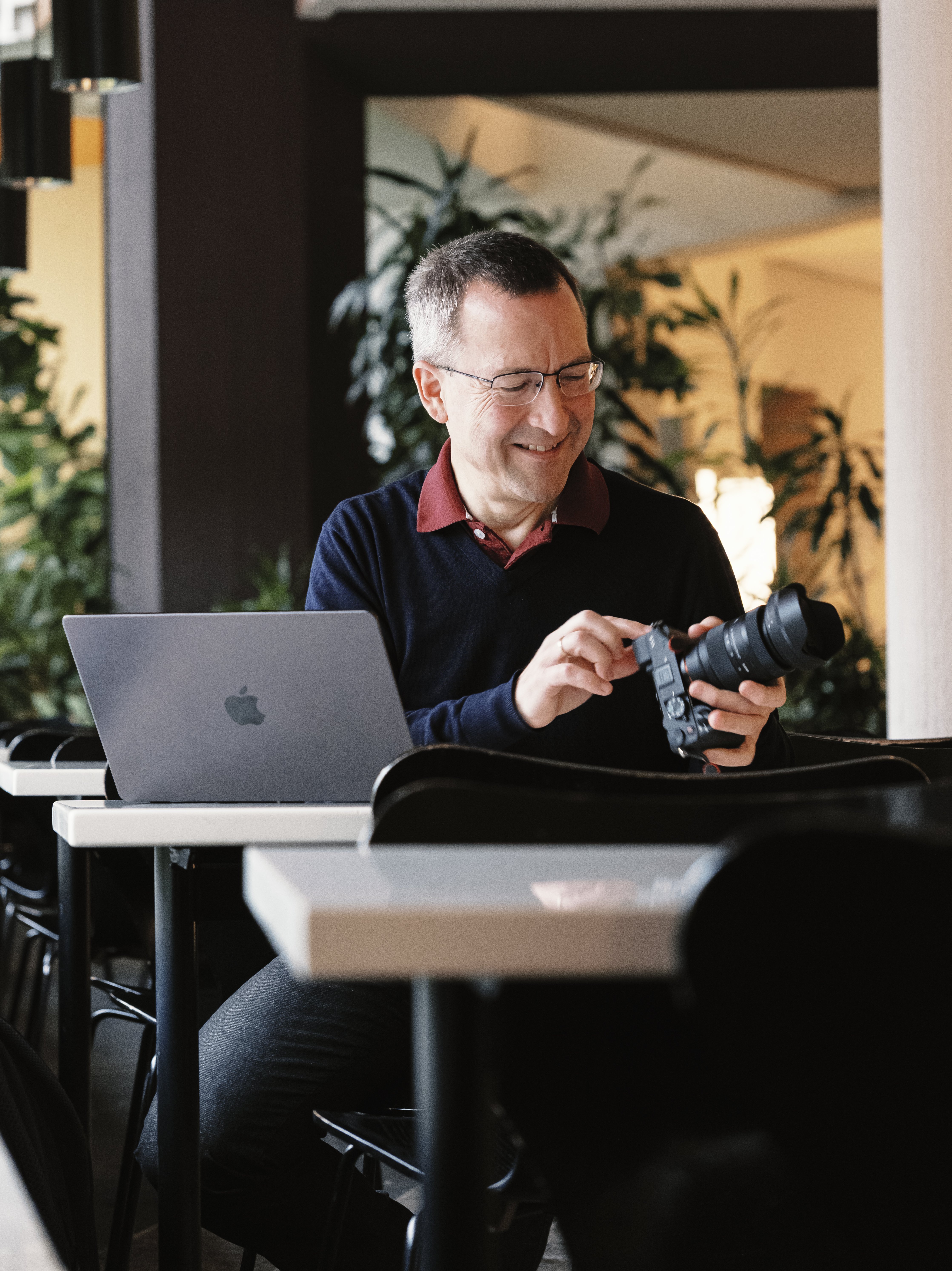 Ein Mann sitzt mit einem Laptop an einem Tisch und lächelt, während er eine Kamera einstellt. Die Kulisse scheint ein moderner Innenraum zu sein, im Hintergrund sind Pflanzen und Hängelampen zu sehen.