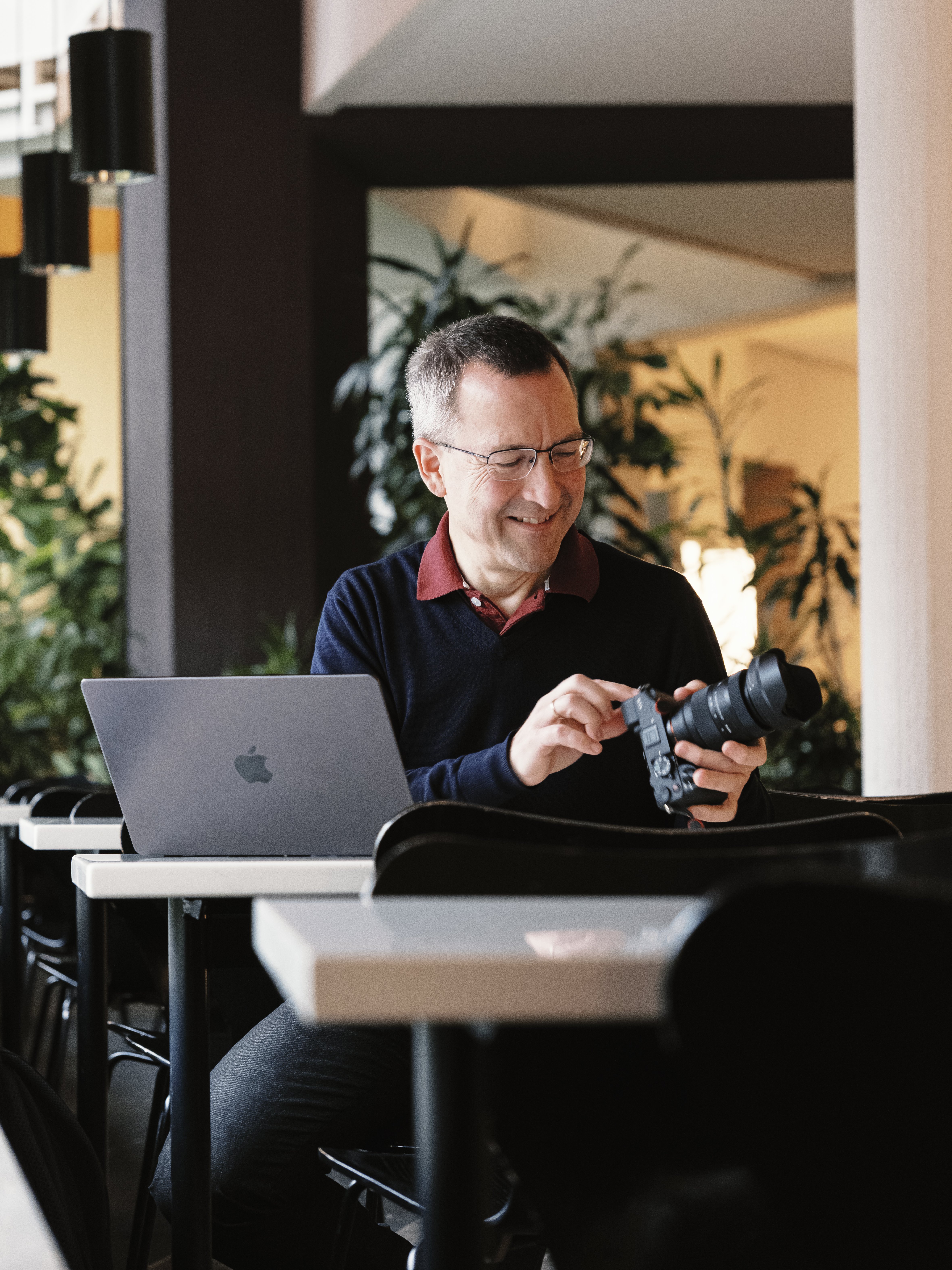 A man sits at a table with a laptop, smiling while adjusting a camera. He is indoors, surrounded by plants and modern decor, with natural light coming from the left.