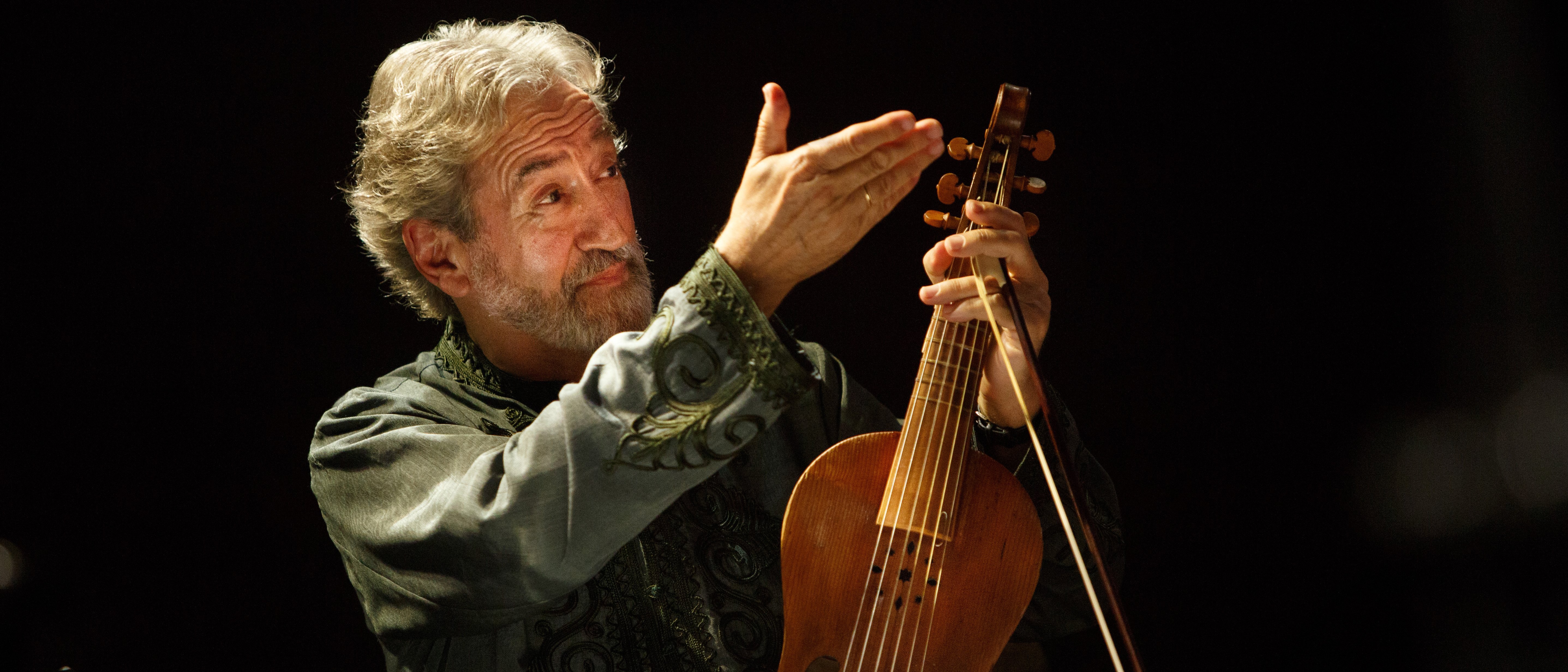 Jordi Savall, with grey hair and a green patterned shirt, holds a wooden stringed instrument in his hand. He is standing on a dimly lit stage. Sheet music and a music stand can be seen in the foreground.